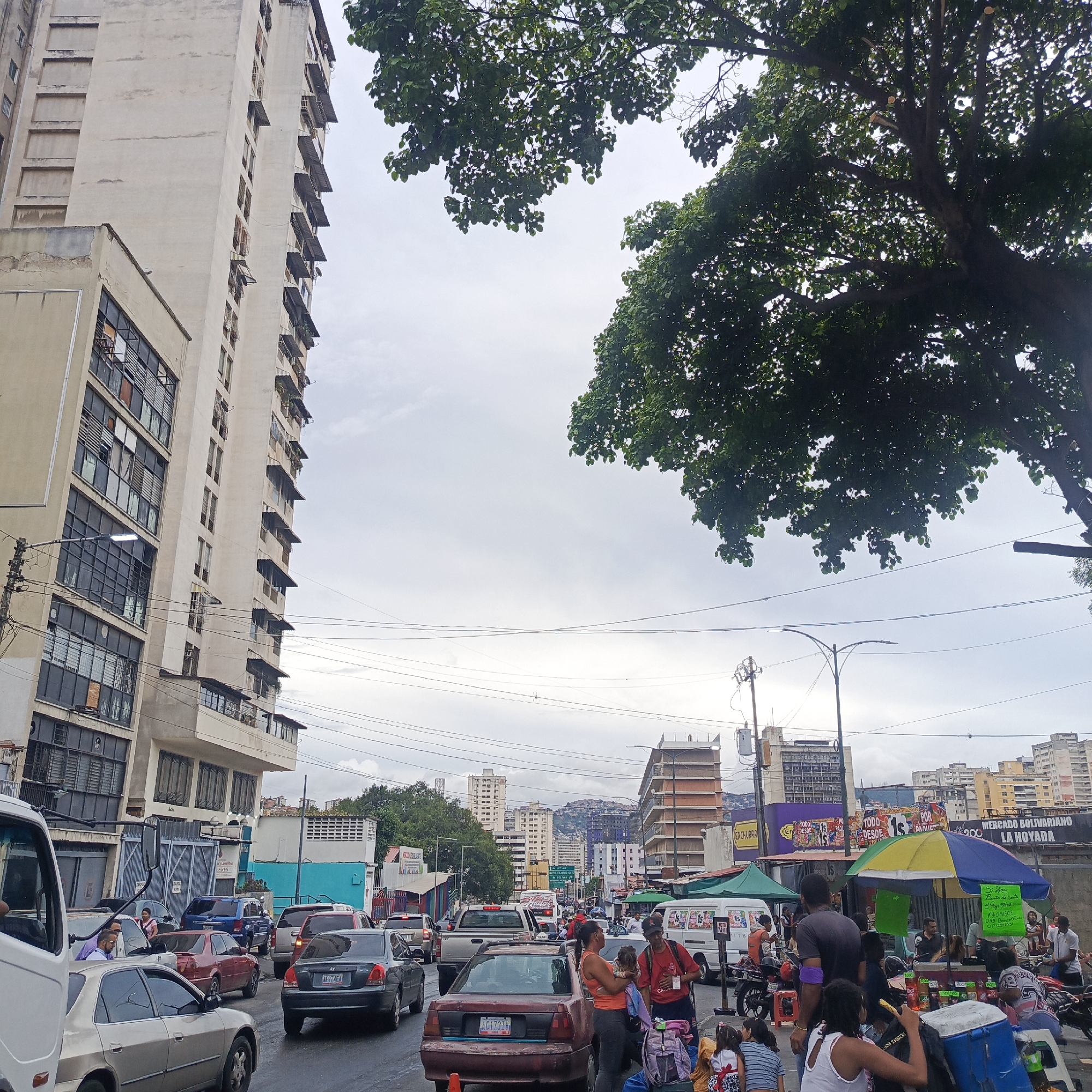 The image depicts a busy street scene in Caracas, Venezuela, likely near the Mercado Bolivariano La Hoyada, given the prominent signage. The environment is characterized by heavy traffic congestion, with numerous cars, vans, and a white truck visible on the wet asphalt road. Tall, light-colored apartment buildings line one side of the street, showing signs of wear and tear, with many windows covered by awnings or grills. On the other side, there are market stalls with colorful umbrellas, food vendors, and various goods displayed. Several people are present, engaged in activities such as shopping, selling, and interacting with each other. Some individuals are standing and talking, while others are seated near the market stalls. A family with children is visible in the foreground, surrounded by bags. Overhead, lush green trees partially obscure the cloudy sky, and a network of power lines crisscrosses the scene, a common sight in urban Venezuelan landscapes. The overall atmosphere suggests a vibrant, albeit chaotic, daily life in a commercial and residential area of Caracas.