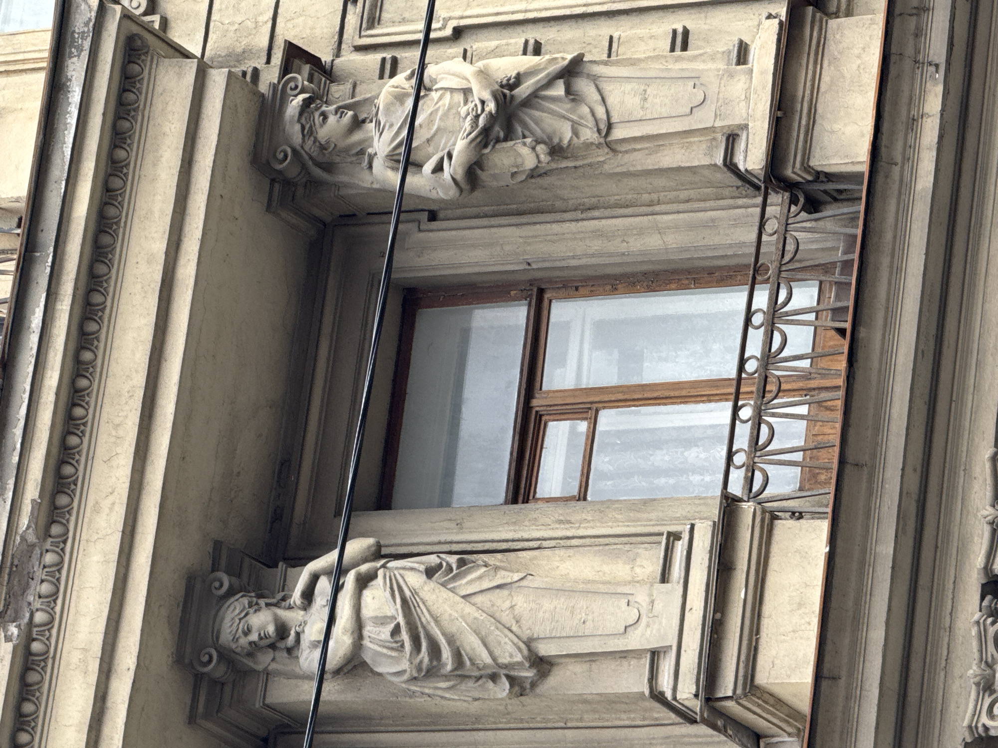 Architectural detail in Saint Petersburg, Russia, featuring a section of a building facade. The frame shows a window with a dark wood frame. Above and below the window are ornate stone sculptures of reclining figures with classical draping. The building's facade is detailed with vertical columns and decorative molding. A metal security grille is positioned outside the window. A wire runs vertically across the scene.