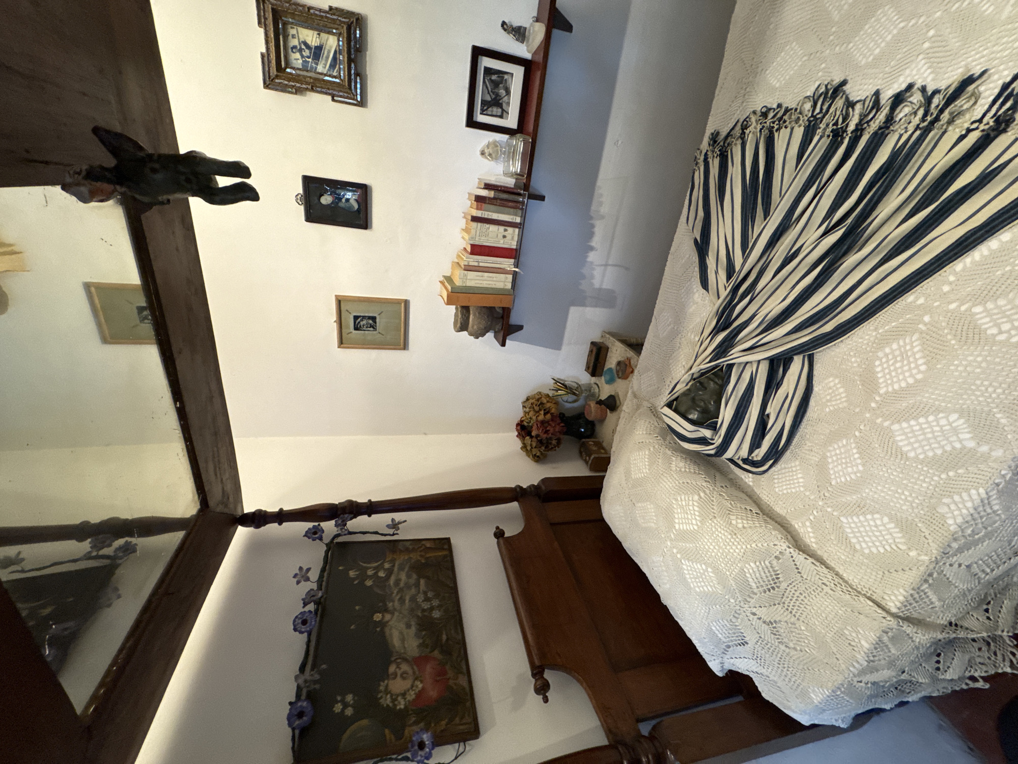 The interior of a bedroom, potentially within Frida Kahlo's Casa Azul (Blue House) in Coyoacán, Mexico City, Mexico. The room has white walls adorned with framed pictures, including portraits and landscapes. A shelf displays a stack of books and small decorative objects. A wooden four-poster bed is partially visible, covered with a white crocheted bedspread and a blue-and-white striped blanket. A painting of a woman is mounted on the wall by the bed. A mirror shows reflections within the room, including an animal sculpture mounted on a beam. The light is soft and diffused.