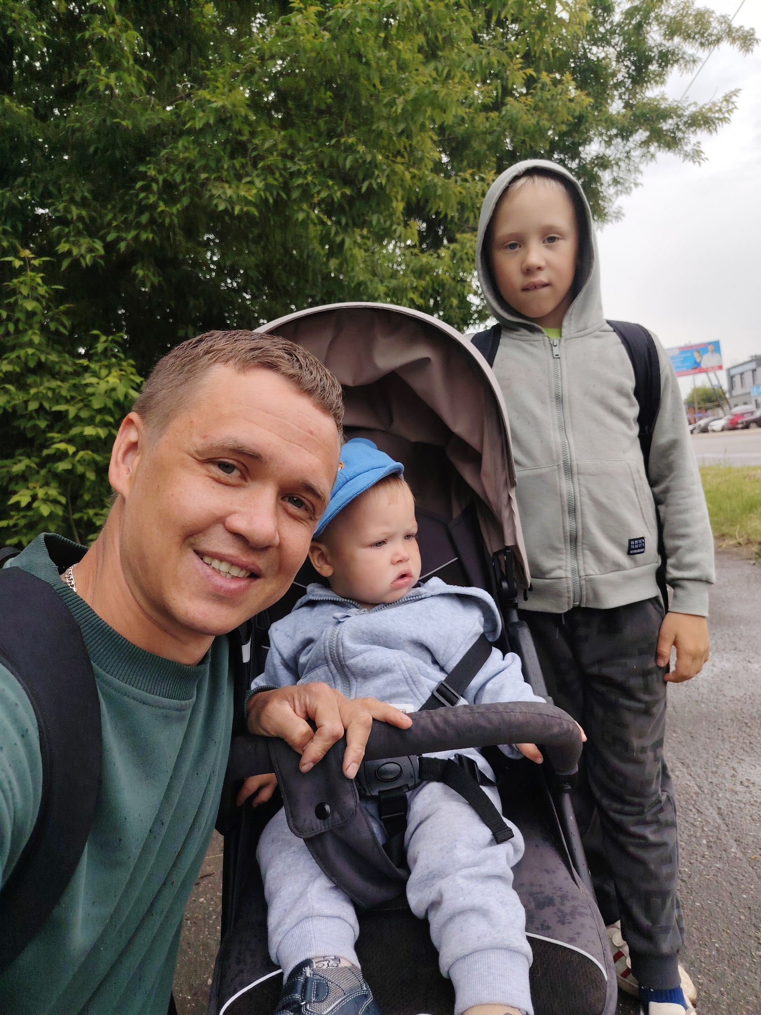 An outdoor selfie captures a father and his two young sons in Chaikovsky, Russia. The adult male, positioned in the foreground on the left, smiles brightly at the camera. He wears a dark green long-sleeved shirt and a black backpack. To his right, a toddler or baby sits in a dark-colored stroller, looking slightly away with a serious expression. This youngest child wears a light grey hooded tracksuit and a blue cap. Standing next to the stroller on the right is an older boy, who looks towards the camera with a neutral expression. He is dressed in a light grey hooded zip-up jacket, dark patterned pants, and a black backpack. On his jacket, a small patch with white text appears to read "30 25 10" and possibly "NEVERLAND".
The family appears to be on a walk, likely on a paved path or sidewalk next to a road, as indicated by the faint presence of cars and buildings in the far background on the right. Lush green trees form a dense backdrop, suggesting a natural or park-like setting adjacent to an urban area. The sky is overcast, providing soft, diffused lighting, and the ground appears slightly damp, hinting at recent rain or high humidity. A billboard with a person's face is partially visible in the distance. The overall mood is casual and familial.