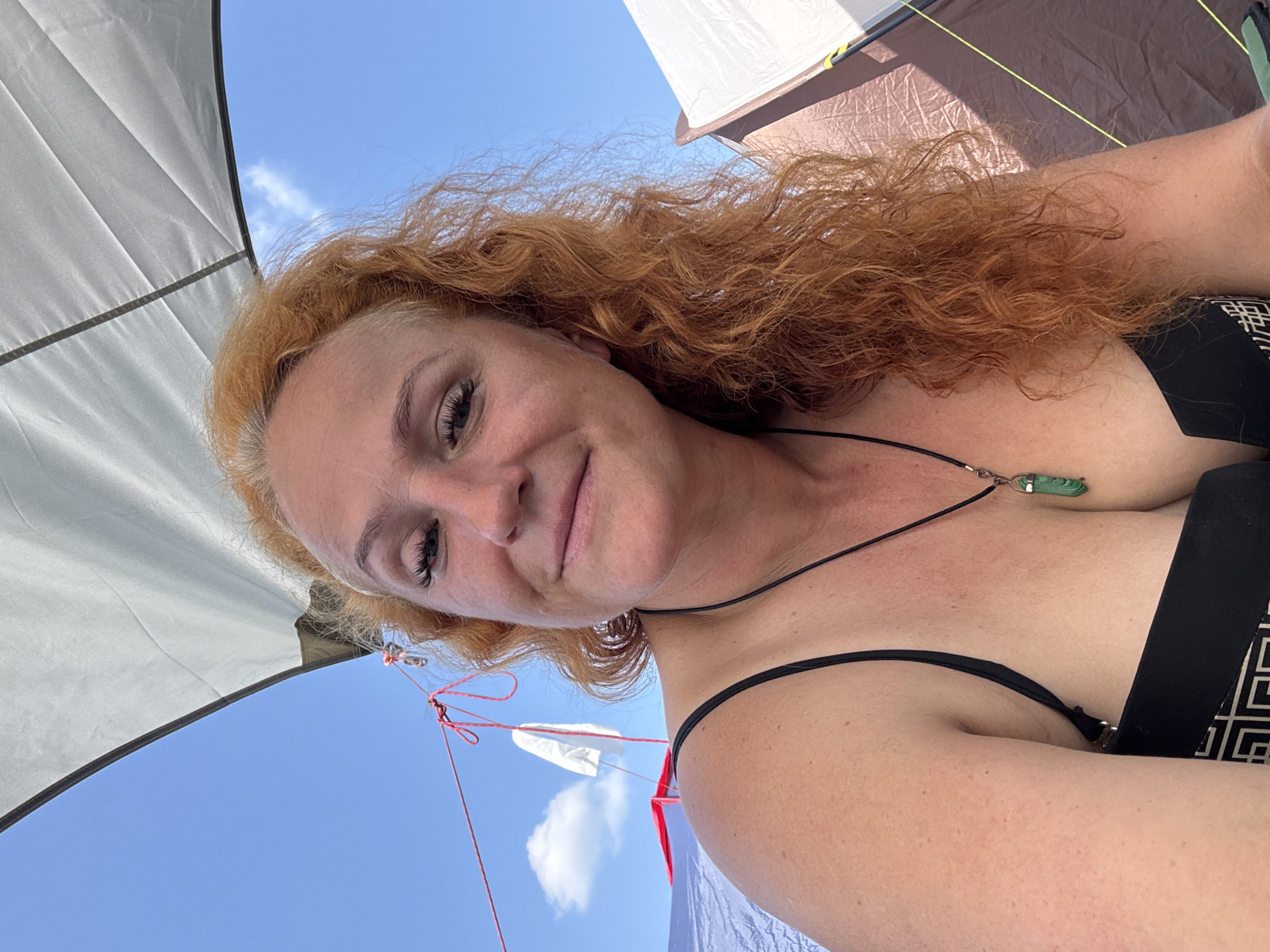 A woman with red, curly hair and a black bikini top is seen from the chest up, outdoors under a bright blue sky. A large white canopy is angled above her, casting a shadow. She is wearing a necklace with a green pendant. In the background, two tents are visible, one blue and red, and another brown and white. A clothesline with a small white object attached stretches across the frame. The woman is smiling gently at the camera.