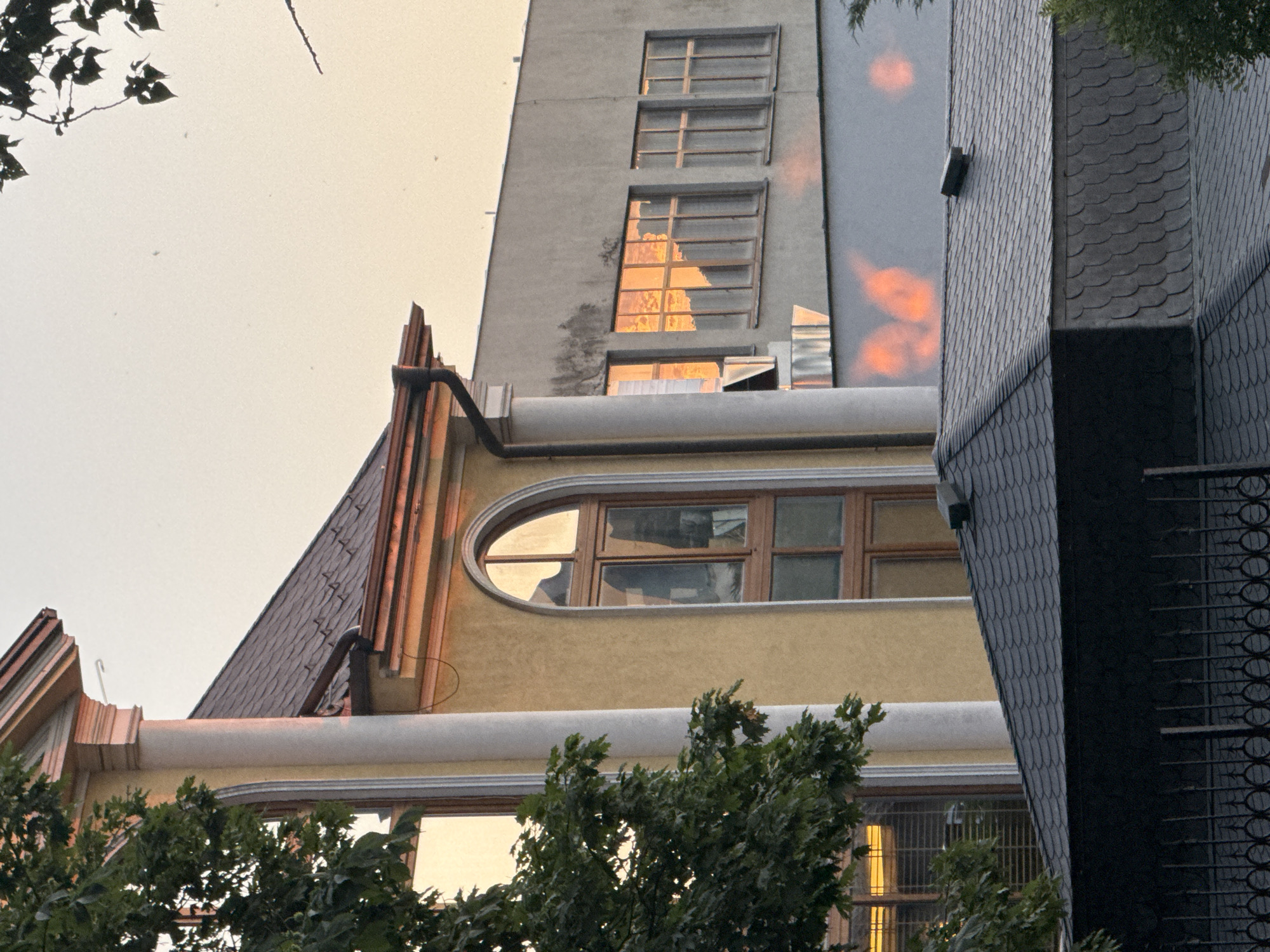 The image shows a close-up, eye-level shot of a building's facade at golden hour, likely in Sofia, Bulgaria, given the context. A large, cream-colored column, part of a classical architectural element, is prominent on the left, partially obscured by the lush green leaves of a tree. To its right, a yellow building features a tall, arched window with wooden frames, reflecting the warm, orange hues of the setting sun. Further to the right, a grey building has multiple windows, also reflecting the sunset, creating bright, abstract patches of light on its surface. In the foreground, a dark grey, shingled roof of a lower structure is visible, with a black metal fence in front of it. The sky is a pale, hazy orange, with a few small specks that might be insects or dust. There are no people visible in the image, and the focus is on the architectural details and the play of light.