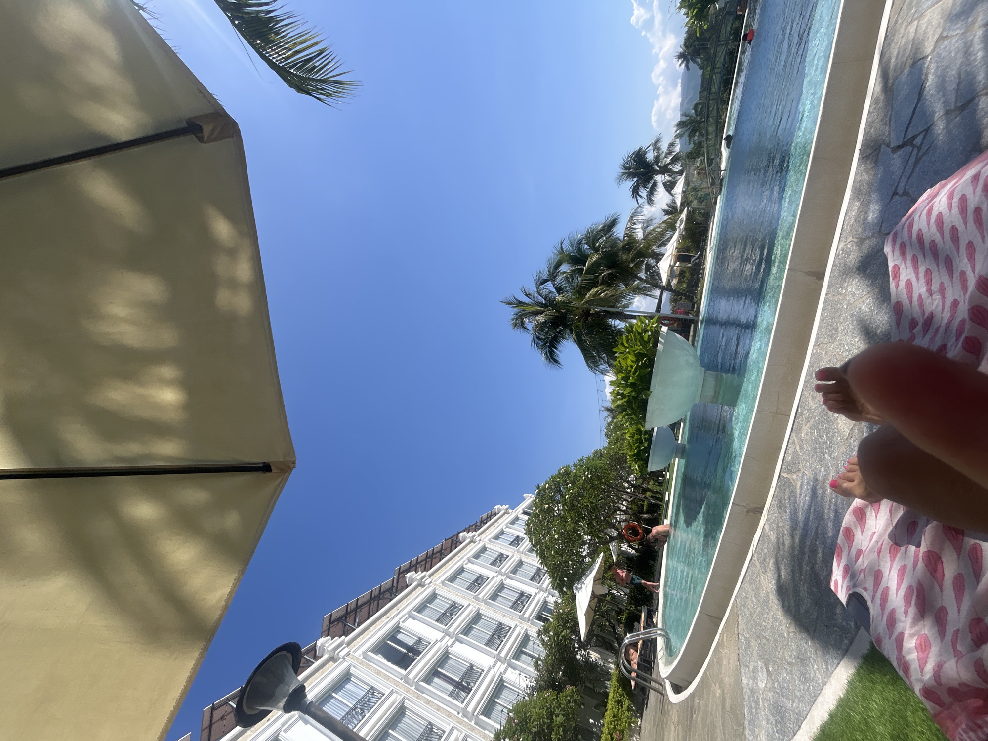 A ground-level, rotated eye-level shot shows a person relaxing near a pool at a resort in Nha Trang, Vietnam. The scene includes parts of a large beige parasol in the lower left, obscuring the view of the clear blue sky with scattered clouds. Palm trees sway in the breeze in the middle of the shot. A long, light-blue pool runs horizontally, with people swimming or standing in the water. A modern white multi-story building is visible in the lower portion of the shot, behind lush green foliage and additional parasols along the pool. In the right corner, the feet of a person with pink painted toes are visible, lying on a pink and white patterned towel. Gray tiling is around the pool, and artificial green turf is in the lower right corner.