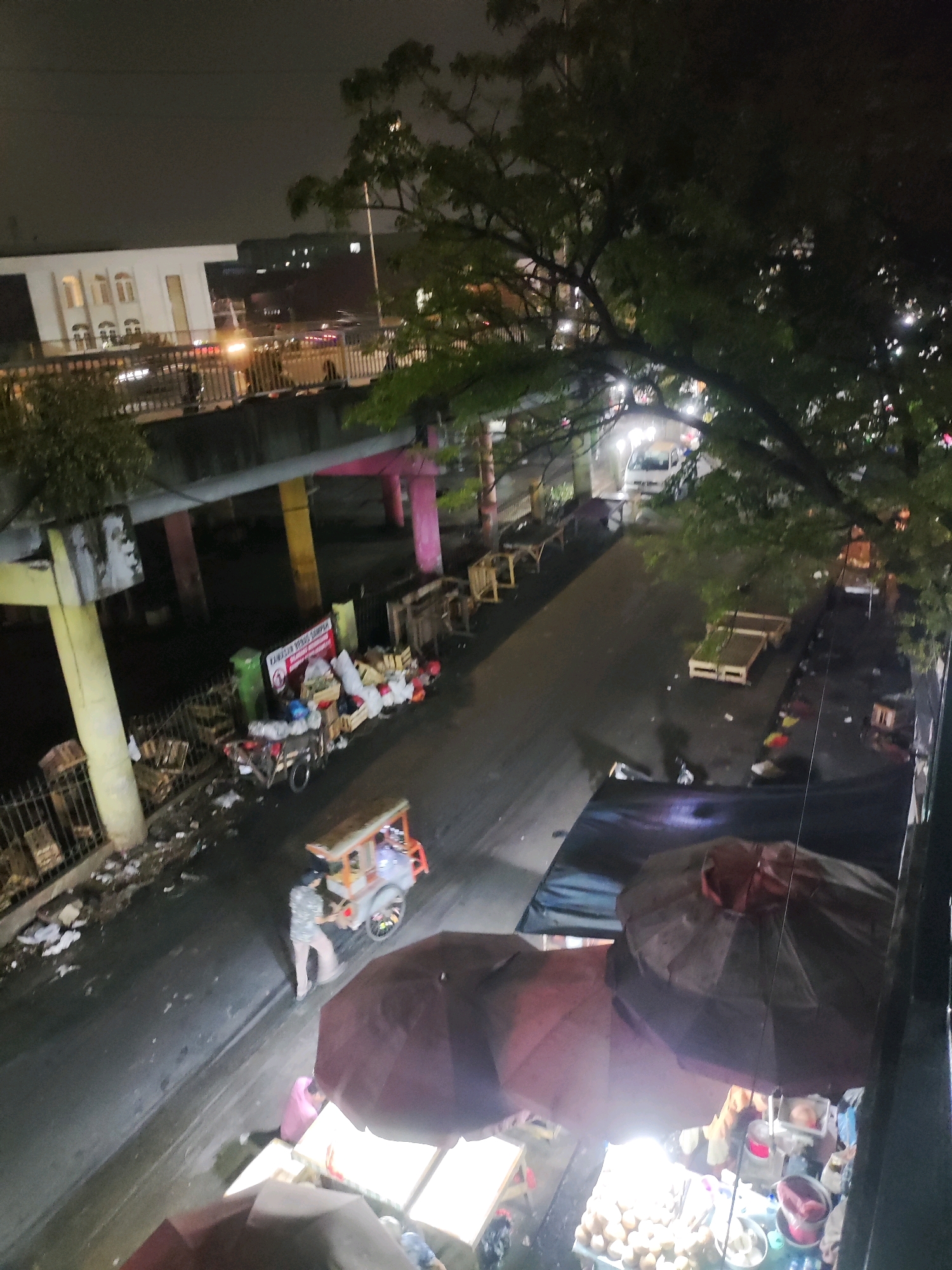 The image captures a nighttime street scene in East Jakarta, Indonesia, characterized by a wet road and overhead illumination from streetlights and vehicle headlights. In the foreground, two large umbrellas, possibly for market stalls, are visible, with one partially open and the other fully open, obscuring some of the scene below. Underneath the open umbrella, various items, including what appear to be food items like eggs and drinks in bottles, are displayed on tables.
Further back, a street vendor operates a cart equipped with lights, and a person in a patterned shirt and light-colored pants is attending to it. To the left of the cart, a motorcycle is parked and loaded with goods, including crates and bags. A green trash can and more wooden crates are also present. In the midground, a road passes under an elevated structure, likely an overpass or flyover, supported by brightly colored pillars (yellow and pink). Vehicles can be seen on the road, with headlights indicating traffic. The area beside the road appears to be a marketplace or storage area with more wooden crates and debris scattered around. In the background, buildings with illuminated windows are visible, suggesting urban development. The overall atmosphere is somewhat chaotic and reflects a busy urban environment at night, possibly during or after rainfall given the wet surface.