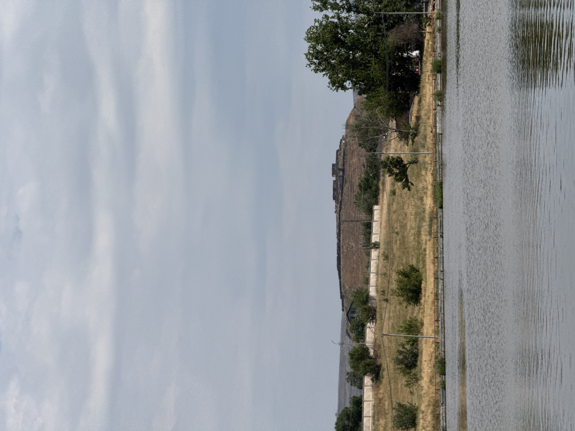 The image shows a body of water in the foreground, possibly a lake or reservoir. Behind the water, there is a grassy embankment lined with a low concrete fence and a red railing. Scattered trees and bushes dot the landscape. In the middle ground, a hill rises with a fortified structure or ruins on its summit. The hill is covered in dry grass and features a building partially visible amongst the trees. The sky is overcast with light clouds. This scene is of the area around the "Lebanese House" and the "Children's Railway" in Yerevan, Armenia, which is situated near a reservoir often referred to as the "Yerevan Sea" or "Abovyan Reservoir."