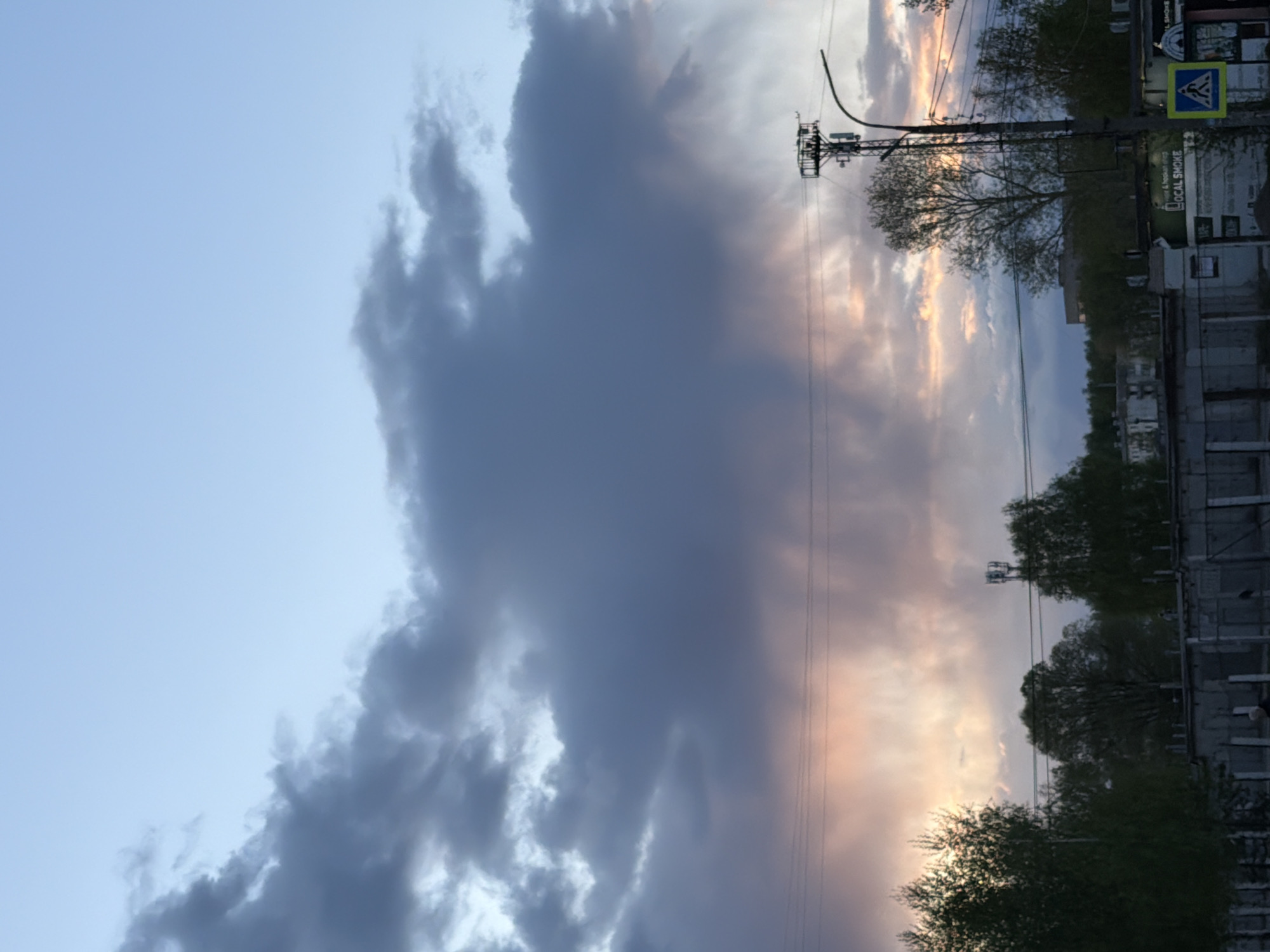 The image presents an evening sky filled with dramatic, dark gray clouds contrasted against lighter, pastel-colored clouds illuminated by the setting sun. Below the sky, on the right side of the frame, is a portion of a cityscape. The buildings appear to be of standard construction, lining the street. Trees are visible along the street, partially obscuring the buildings. Utility poles and wires run along the street. A pedestrian crossing sign is visible in the foreground. The text “LOCAL SMOKE” is partially visible on the street. Given the location of the image, it is possibly somewhere in the city of Chaikovsky, Russia.