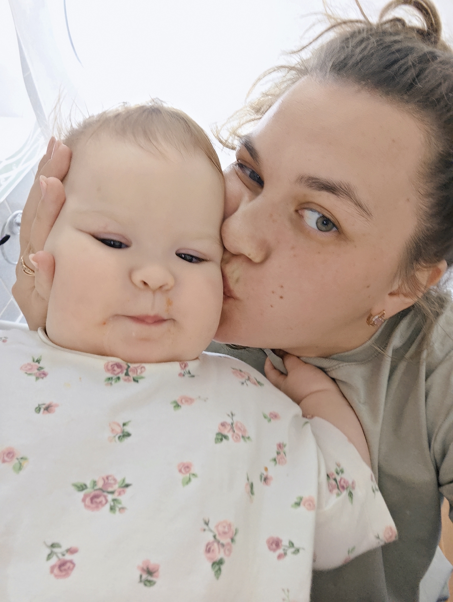 A light-skinned woman with her hair pulled back is kissing a baby with similar complexion on the cheek. The baby is wearing a white top with a floral pattern. The woman is wearing a green or grey top. The scene is indoors, possibly in a home with white curtains or blinds visible in the background. The location is unspecified, but the user mentions the city of Chaikovsky, Russia.