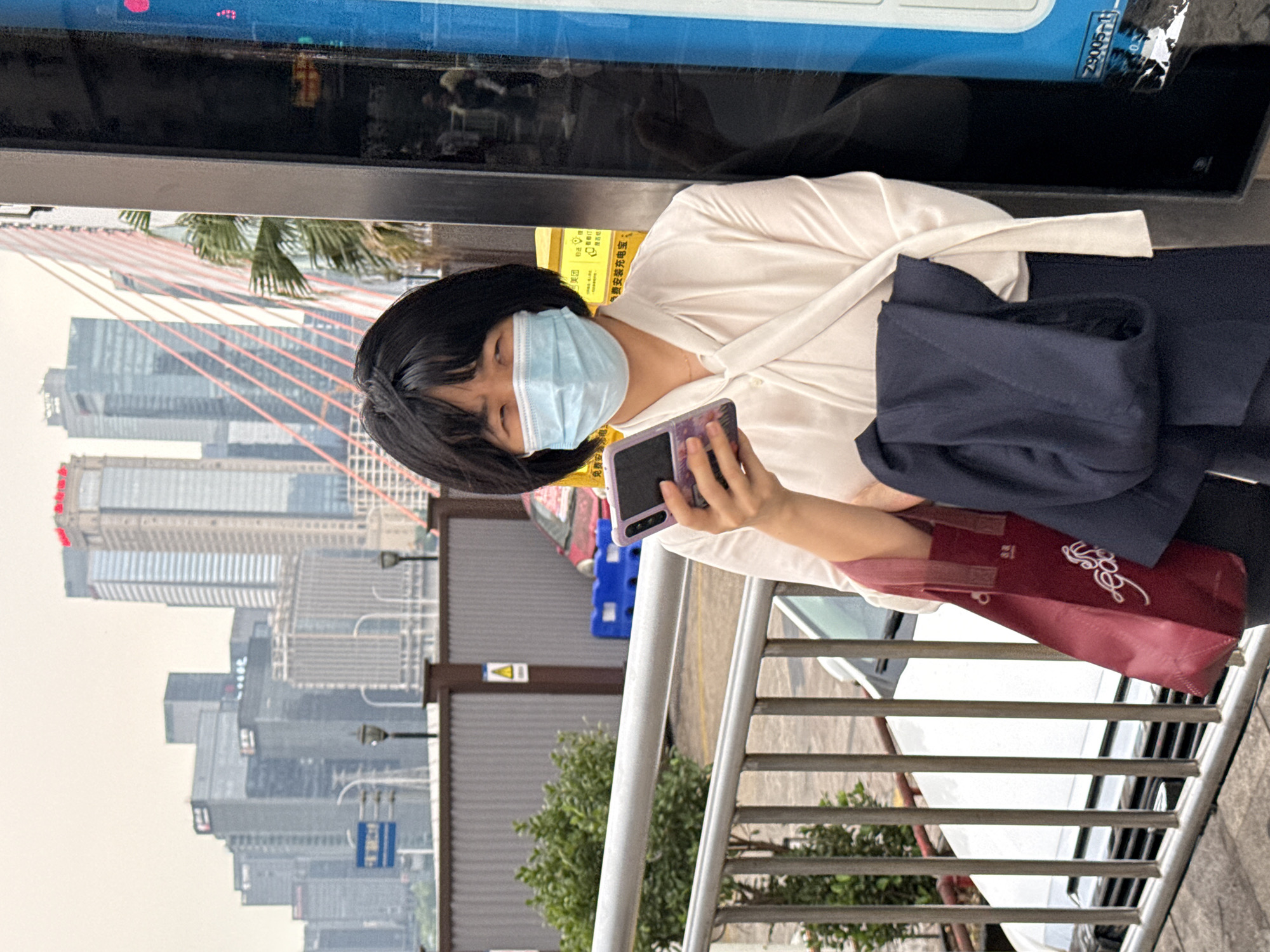 A woman wearing a white blouse and a light blue face mask is holding a purple smartphone and a burgundy shopping bag. She is standing next to a metal railing, with a cityscape featuring tall buildings and a bridge visible in the background. The location appears to be Yuzhong District, Chongqing, China, based on the architecture and the bridge visible in the background, which resembles the Raffles City Chongqing complex. A bus or train is also visible.