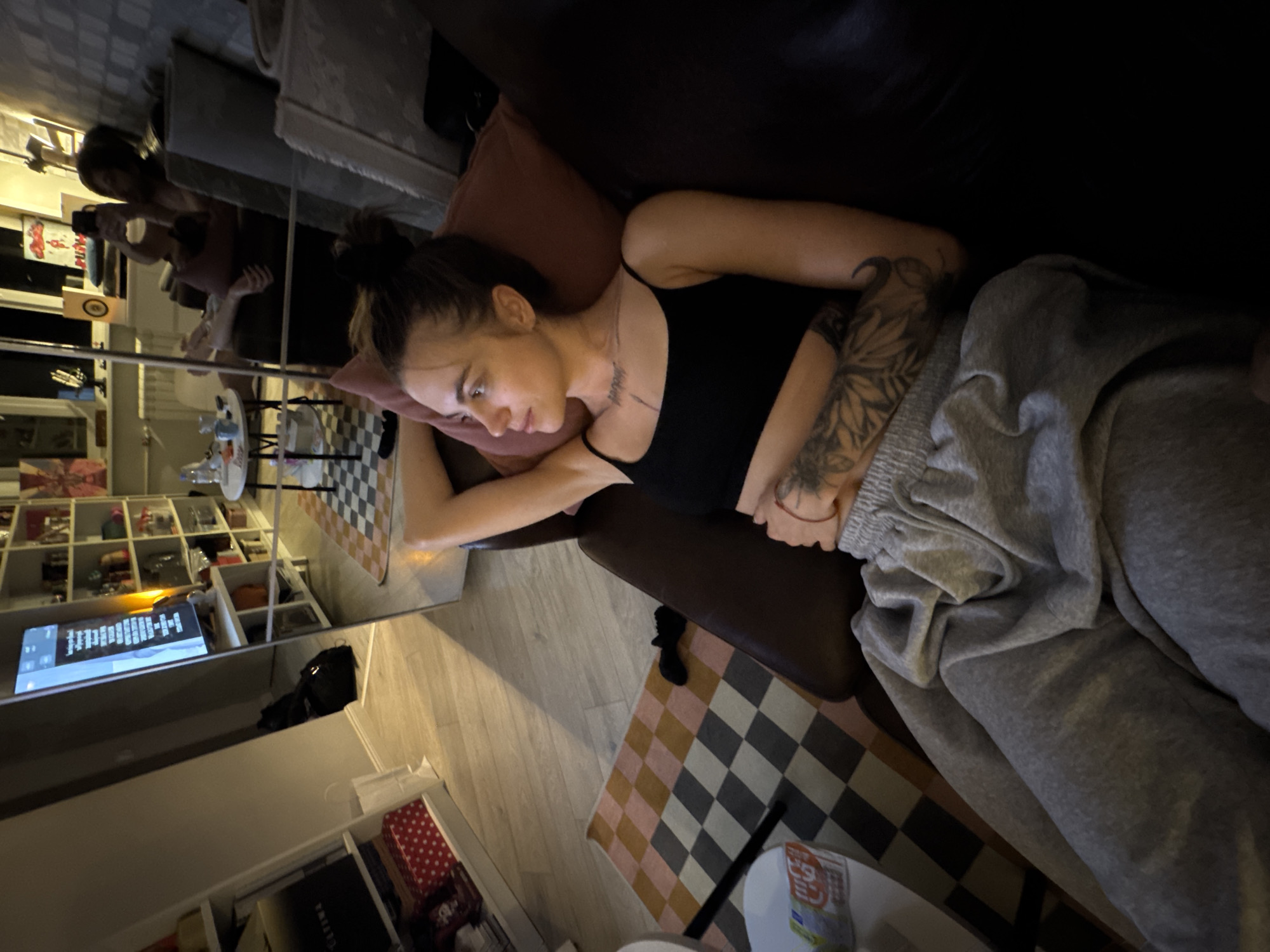 The image shows a young woman lying on a couch in a dimly lit living room. She is wearing a black tank top and gray sweatpants. She has tattoos on her arms and chest. The room appears to be furnished with a couch, a coffee table, and shelves filled with books and decorative items. A television is visible in the background, reflecting the room. The overall impression is of a relaxed, home environment. There is no information within the image to indicate the location is Moscow, Russia.