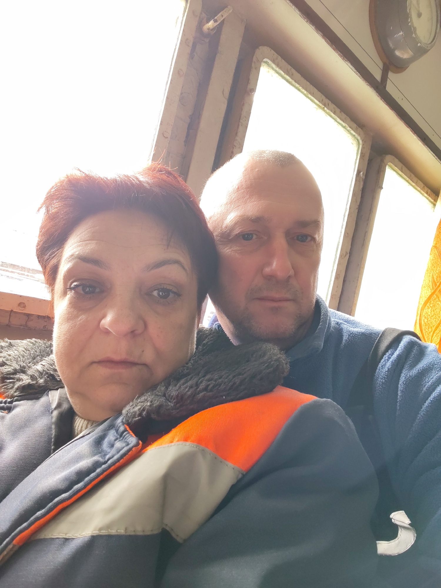 A selfie shows a middle-aged woman and a middle-aged bald man inside a boat or vessel. The woman has short red hair and wears a dark jacket with an orange and gray stripe and a furry collar. The man wears a blue fleece jacket. The background consists of a window frame and a clock. Given the image details and the stated location of Rybinsk, Russia, which is a city on the Volga River, the setting is likely a river transport vessel operating in the Rybinsk area.