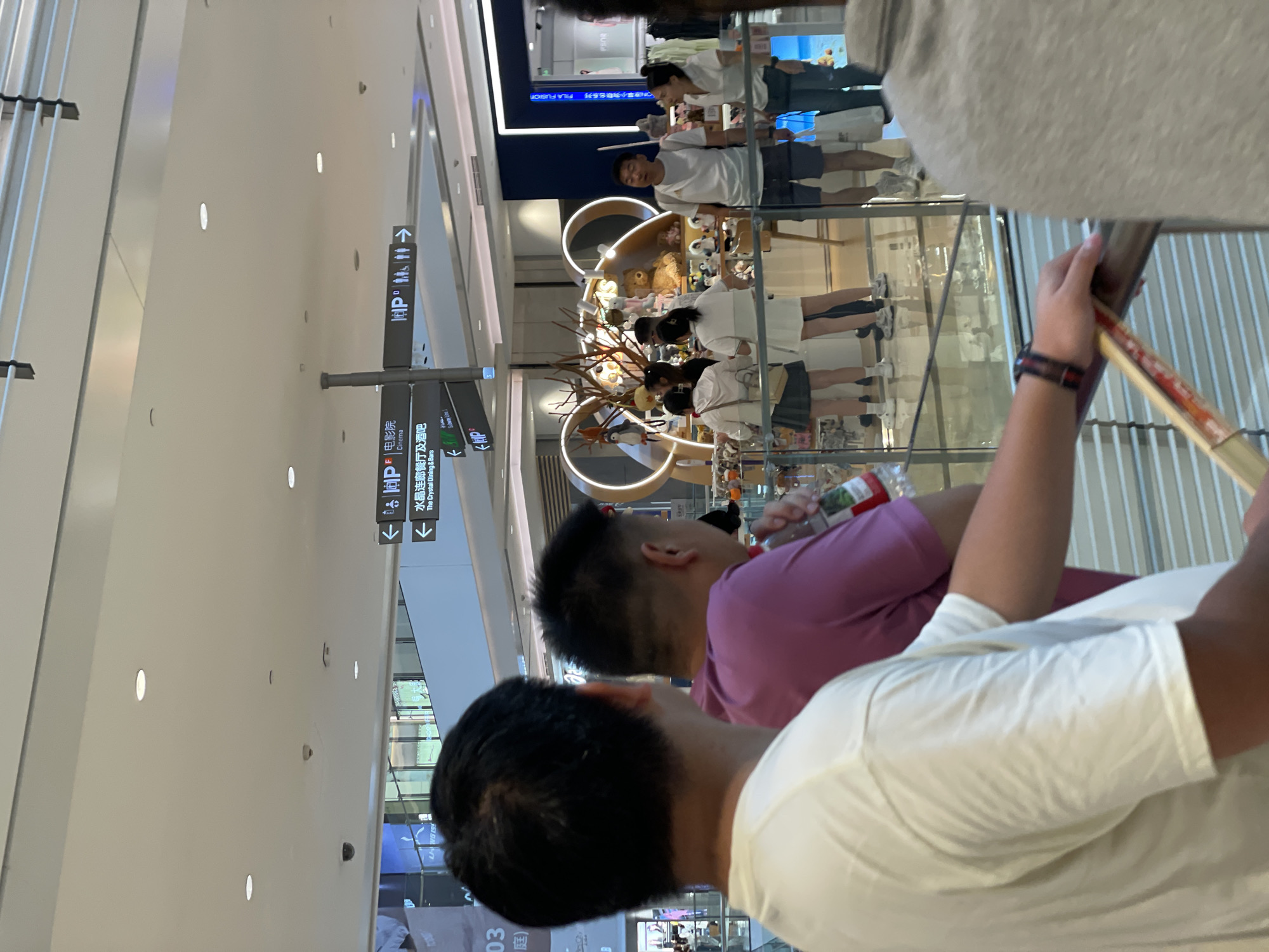 In Yuzhong District, China, the image captures a busy interior scene within a shopping mall. The foreground features the backs of two men, one in a white t-shirt and the other in a purple t-shirt, both facing away from the camera and observing the activity ahead. The man in the purple shirt is holding a bottle of drink. Beyond them, a group of people are gathered around a display, which appears to be a shop selling plush toys. Some individuals are standing, looking at the merchandise, while others are walking past. Directional signs with Chinese characters and pictograms are visible on a pole, indicating "Cinema" and "The Crystal Dining & Bars" with arrows pointing to different directions. The environment is well-lit, with a modern ceiling and reflective flooring. The overall impression is that of a typical commercial setting with shoppers and retail displays.