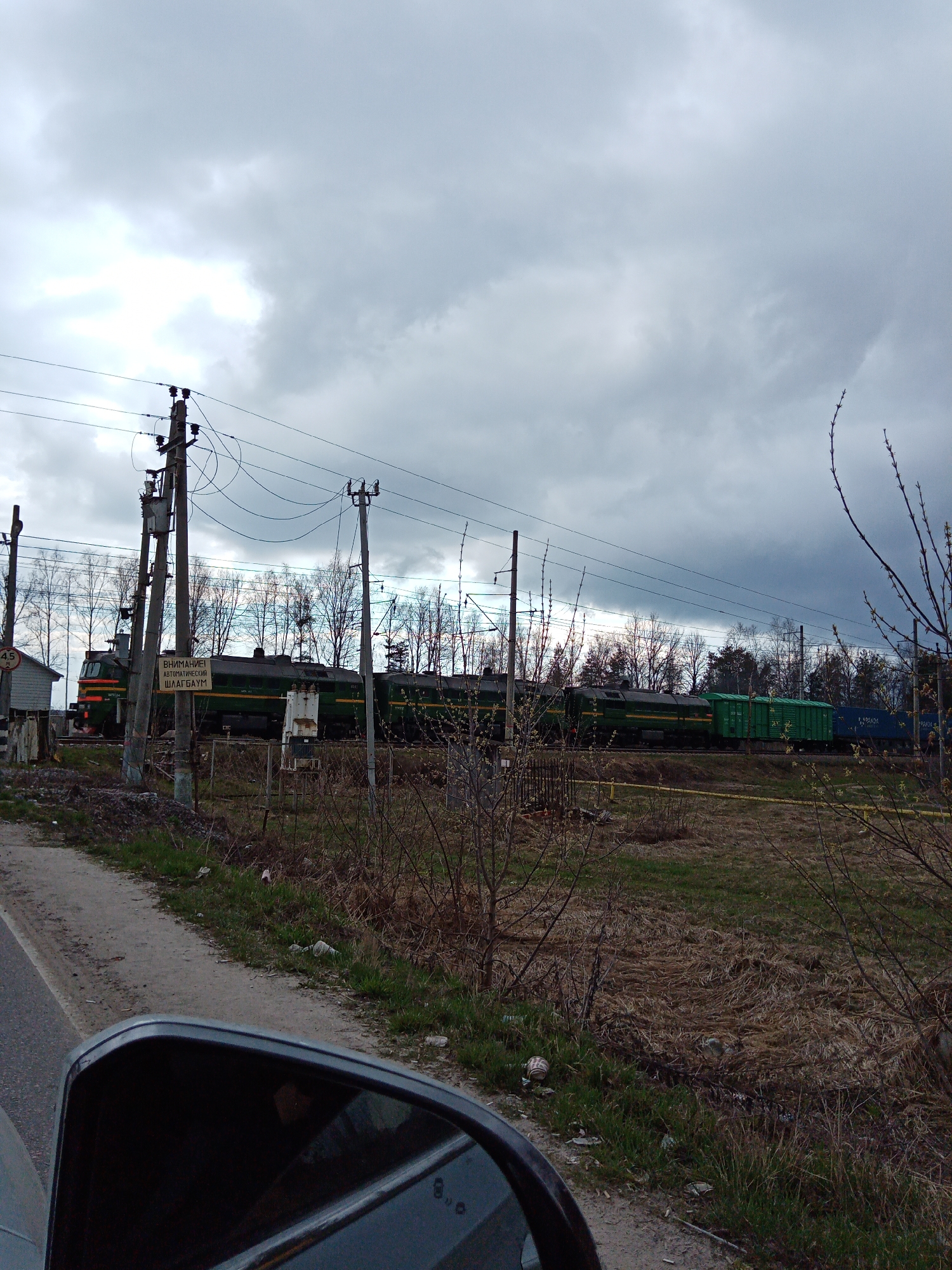 The image depicts a freight train passing through a railway crossing in Kriulino, Russia, under a heavily overcast sky. In the immediate foreground, the dark frame of a car's side mirror is visible on the bottom left, reflecting a dark interior. A dirt path runs alongside dry grass, sparse vegetation, and bare branches, with scattered litter evident on the ground.
In the midground, several utility poles with numerous crisscrossing wires stand prominently. Attached to one pole is a rectangular sign with Cyrillic text: "ВНИМАНИЕ! АВТОМАТИЧЕСКИЙ ШЛАГБАУМ," which translates to "ATTENTION! AUTOMATIC BARRIER." A long, dark green freight train, led by a locomotive, moves across the scene, followed by several green and blue freight cars. One blue container shows partial text resembling "KAPENDA." Behind the train and utility poles, a line of bare trees, characteristic of late winter or early spring, stretches across the horizon. A small, light-colored wooden structure is partly visible on the far left, and a signal box stands near the tracks.
The background is dominated by a thick, grey, cloudy sky, indicating an overcast day. The overall impression is one of a functional, semi-rural landscape, with the industrial elements of the railway and power lines contrasting with the muted natural surroundings.