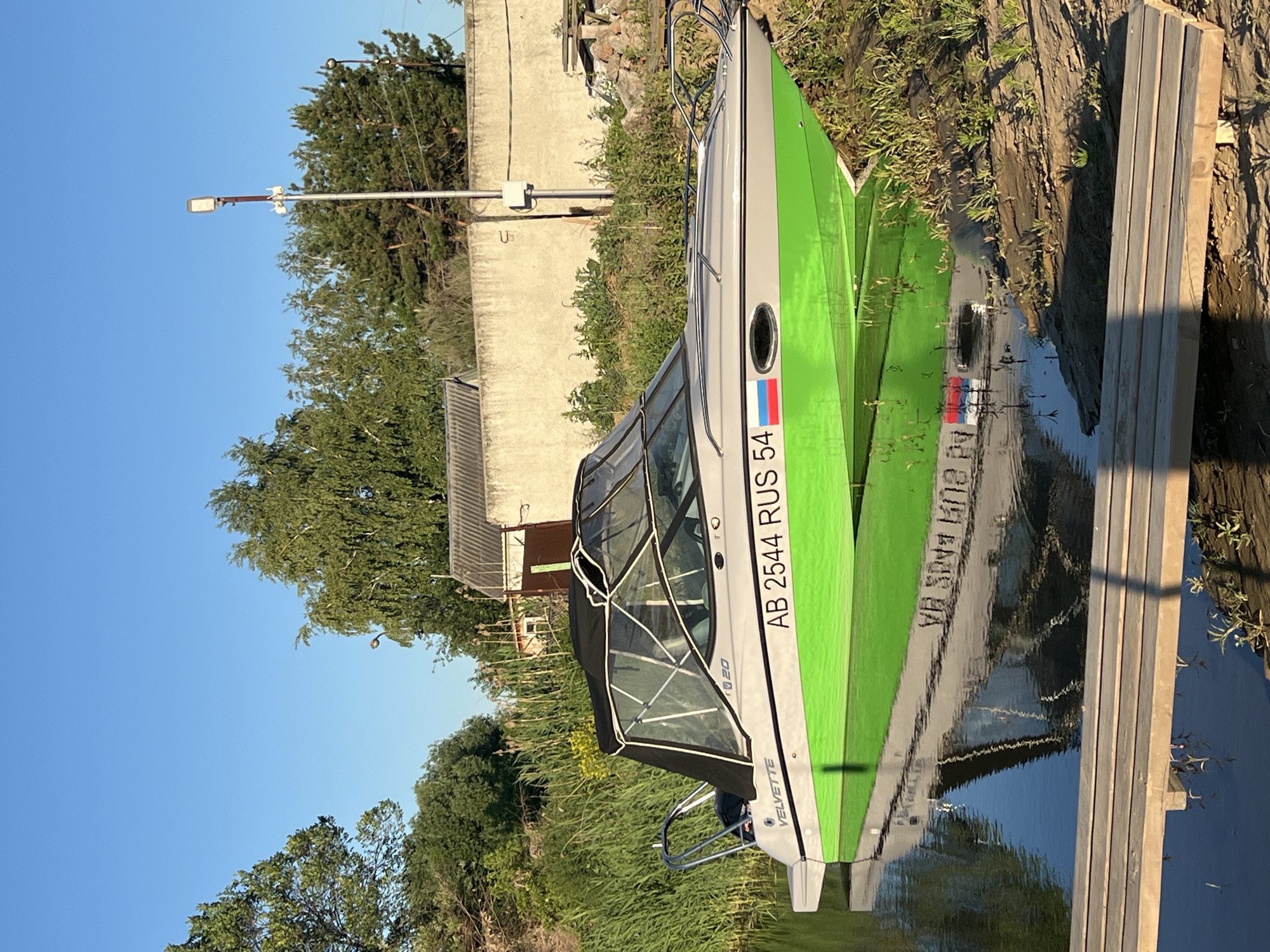A gray and green motorboat with a black canopy is docked on the edge of a body of water, with its hull partially submerged in the water. The boat has the registration number "AB 2544 RUS 54" and a Russian flag sticker on its side. The boat is positioned near a sandy bank covered in dry grass and small plants, with a wooden boardwalk in the foreground. In the background, a concrete wall runs horizontally, with a street light pole attached to it. Beyond the wall, there are trees and vegetation, and a small building with a gray, corrugated roof. The sky is clear and blue, indicating a sunny day. The scene appears to be in Novopichugovo, Russia. There are no people visible in the image.