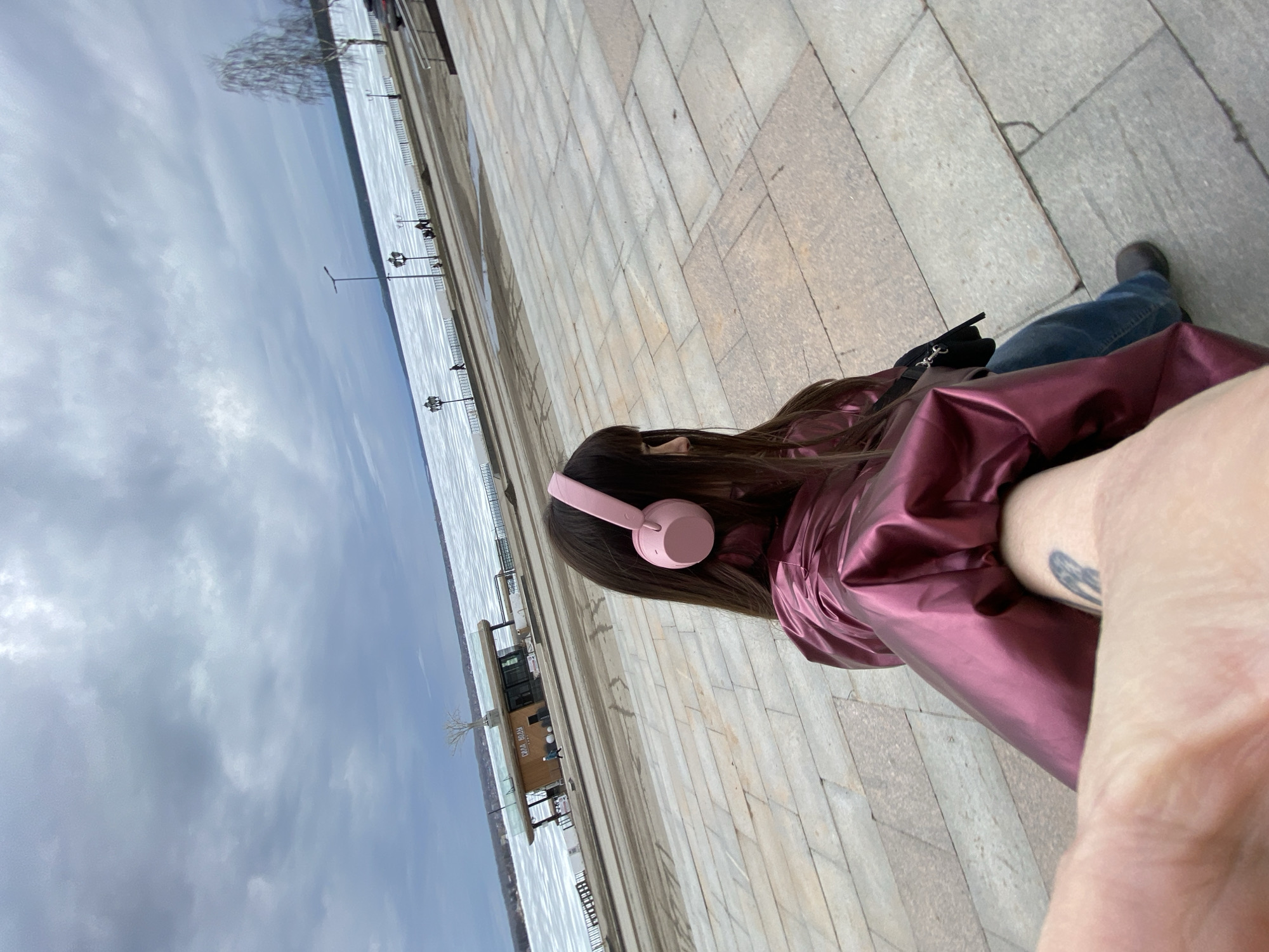 A woman wearing pink headphones and a shiny, maroon-colored coat is seen from an oblique high-angle perspective while standing on a paved surface. The paving consists of rectangular stone blocks. She is facing a body of water, partially covered in ice, along which a promenade stretches with periodic lampposts. Buildings are visible along the waterfront. The sky is overcast with grey clouds. The photo appears to be taken in Votkinsk, Russia, based on the context of the prompt. The woman is wearing blue jeans and black footwear. A black bag is near her feet. The arm holding the camera is visible in the foreground, bearing a tattoo.