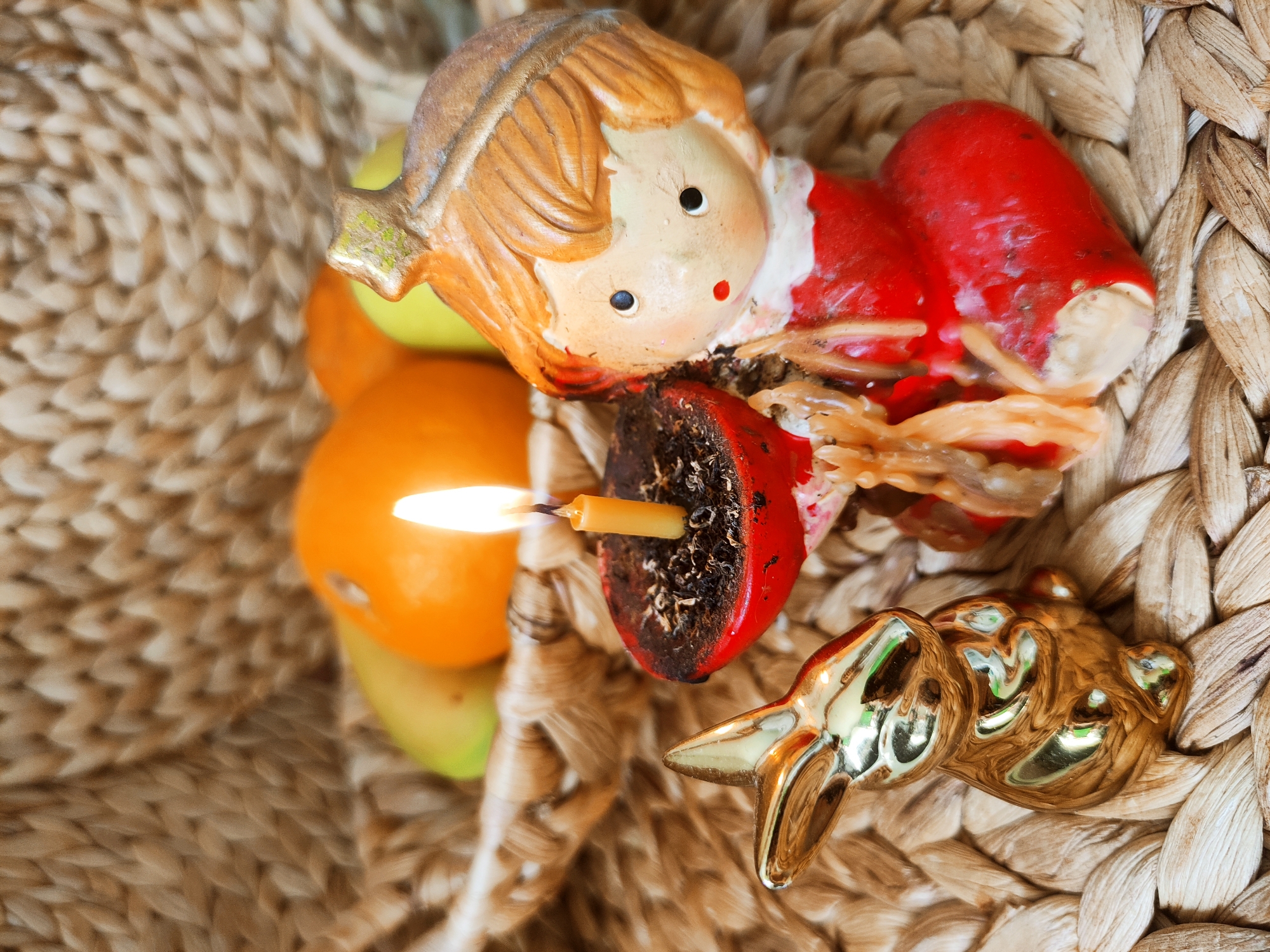 A close-up shot depicts a variety of objects arranged inside a woven basket. Central to the arrangement is a painted ceramic angel figurine, featuring a red robe and a small red dot representing its mouth. To the lower-right of the angel, a gold-colored metal ornament in the shape of a bear is visible. A small candle, still burning with a visible flame, is positioned in a red holder with dark, charred remnants of previous use. Several fruits, including an orange and a green apple, are also included in the arrangement. The entire scene is set against the backdrop of the woven basket. The image does not provide any information about location or time.