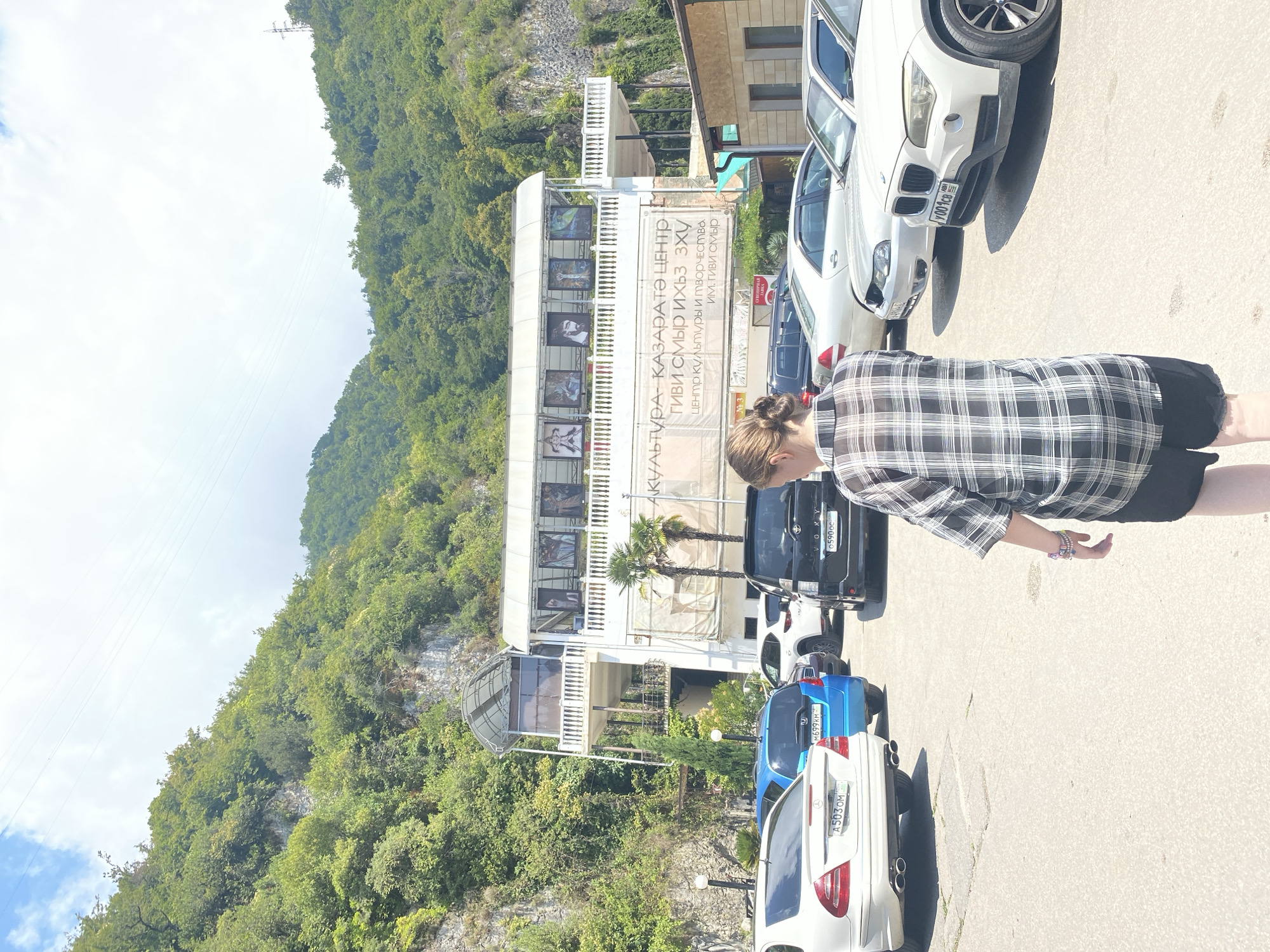 An outdoor, medium shot captures a paved parking lot with several vehicles and a multi-story building situated against a lush green hillside. The building, painted white, features multiple levels, a covered upper balcony with railings, and a large banner displaying text in Cyrillic. Artwork is visible on the upper level of the building. A woman wearing a black and white checkered shirt and black shorts stands in the foreground, facing away from the camera and toward the building. Various cars, including a white Mercedes-Benz, a blue Honda, a black SUV, a silver sedan, and a white BMW SUV, are parked in the lot. The environment appears to be a sunny day with a partly cloudy sky.