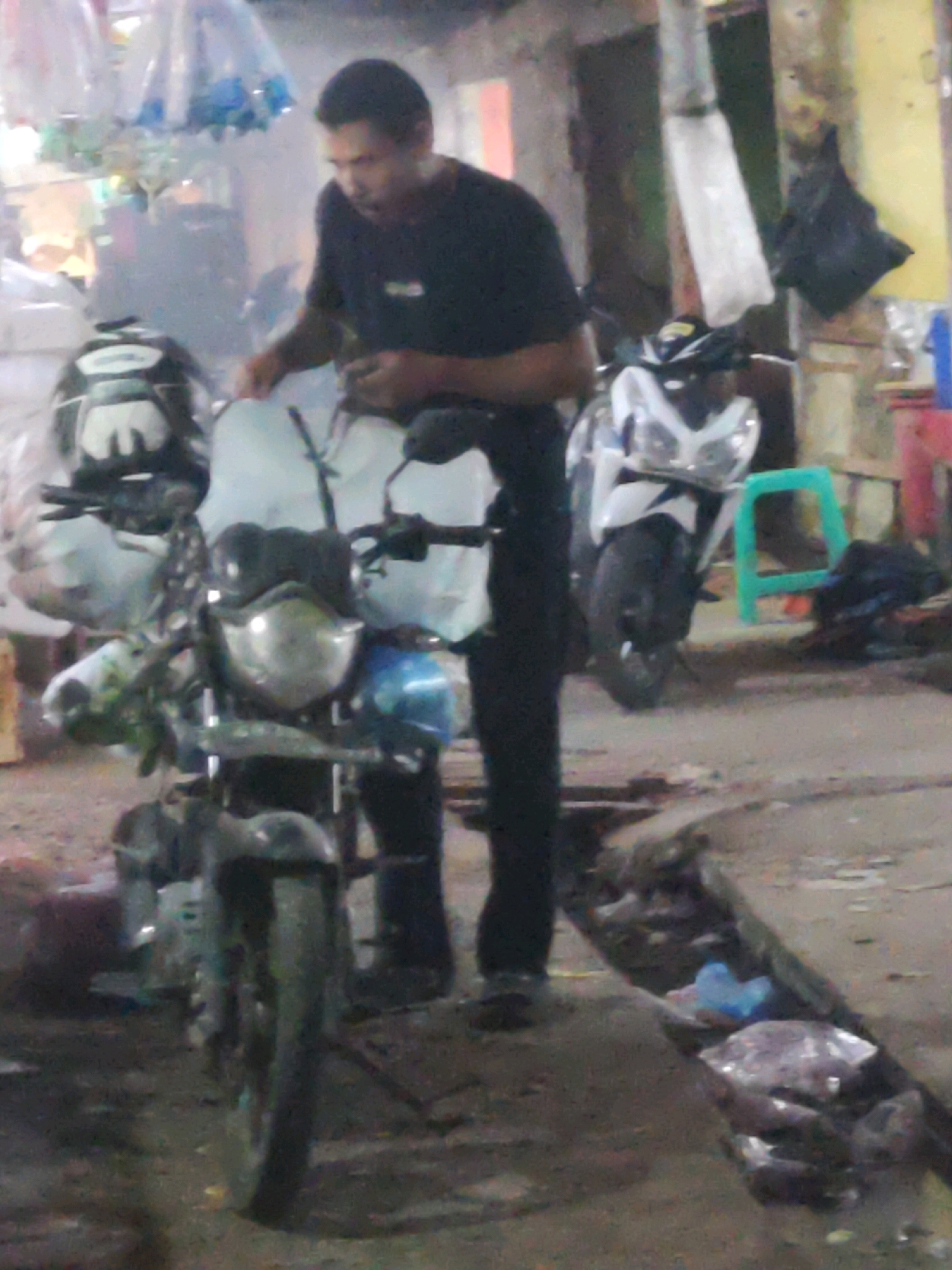 The image depicts an outdoor scene with two motorcycles and a person. A man wearing a dark t-shirt and dark pants is standing next to a motorcycle, appearing to interact with it. In the background, another motorcycle is parked. The environment suggests a street or roadside setting, possibly in a commercial or public area, with some structures and debris visible. The lighting indicates it could be evening or a dimly lit area. The scene is in Karawang, Indonesia, as stated.