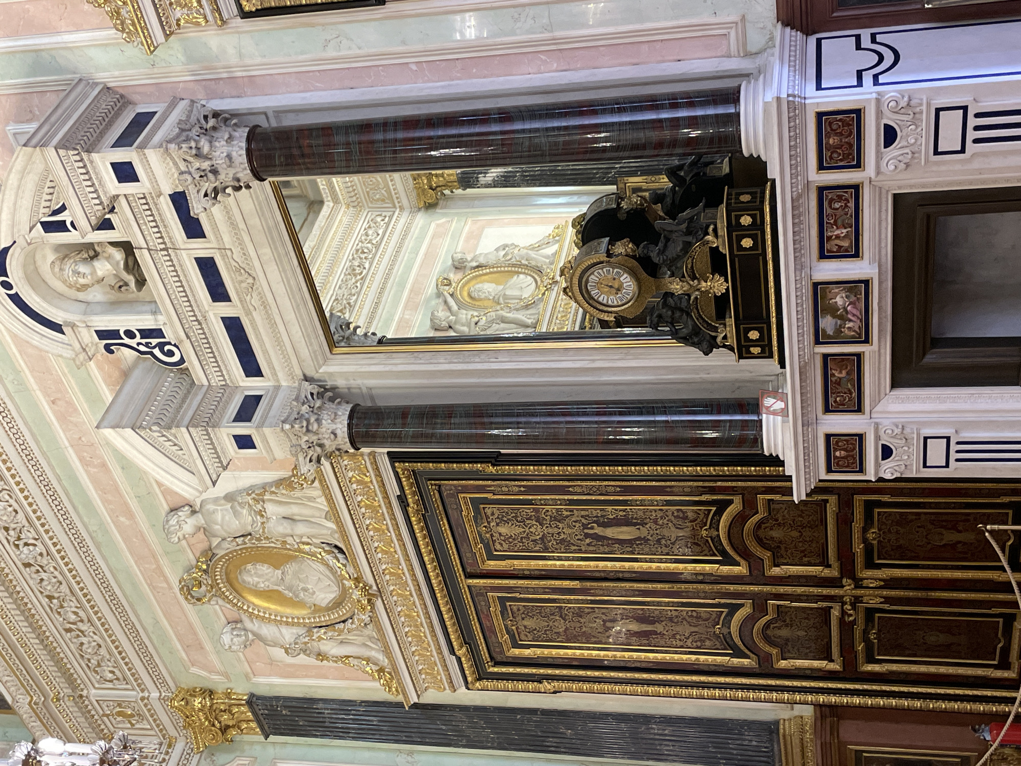 The image depicts an ornate interior, likely a room within a historical building in an unknown city. The scene is characterized by its rich decorative elements.
The walls and ceiling are adorned with intricate stucco work, including sculpted figures and gilded details. A prominent feature is a large dark-colored wooden cabinet or closet, its panels decorated with gold leaf and possibly painted scenes. Adjacent to the cabinet is a framed mirror, reflecting more of the room's ornate ceiling. There is a clock visible above the mirror. The architecture incorporates classical elements like columns with Corinthian capitals. The overall style suggests Baroque or a similar period of elaborate European design.