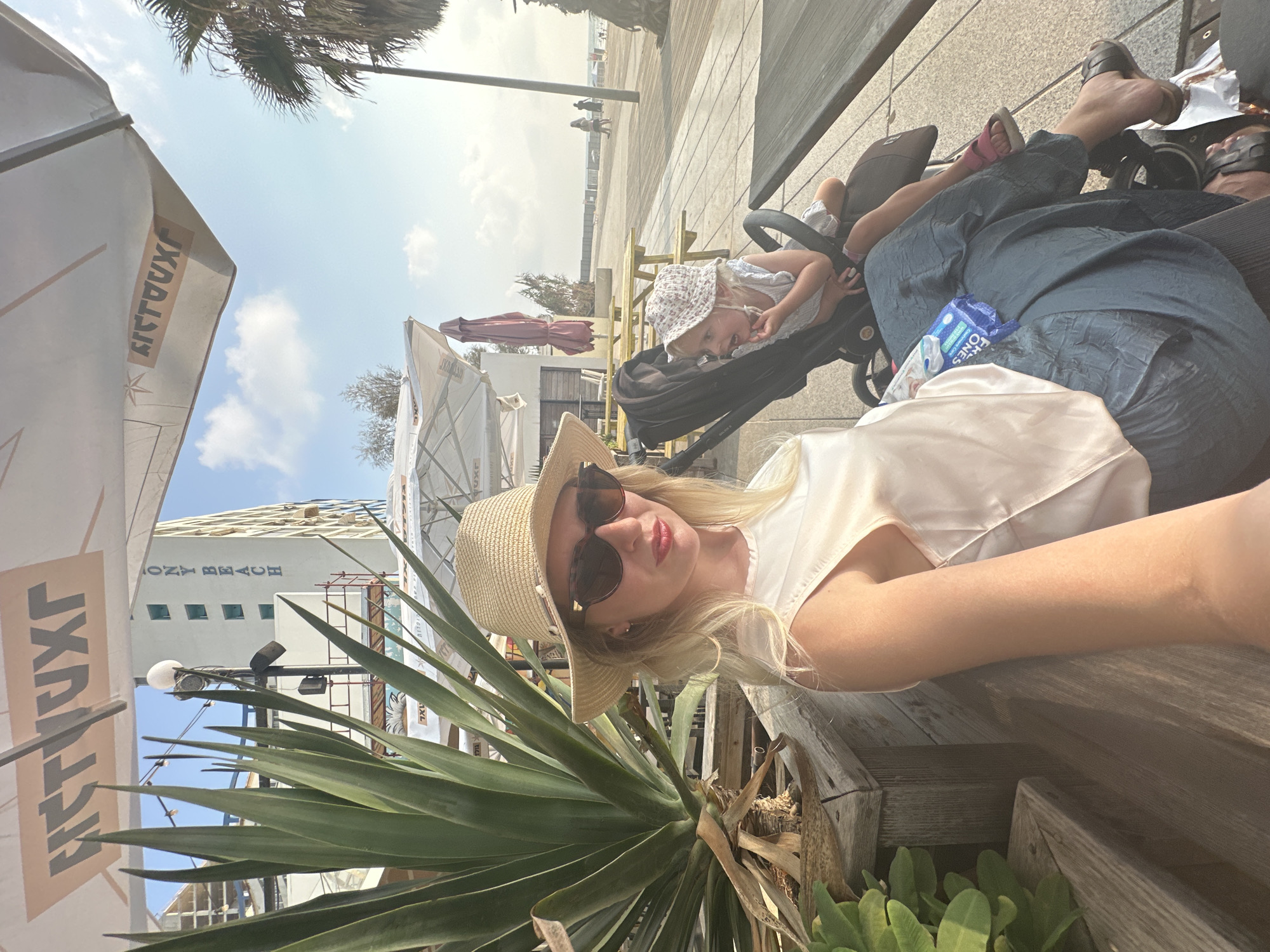 This is a selfie taken by a woman sitting outdoors at what appears to be a beachfront cafe in Bat Yam, Israel. The woman is wearing a straw hat and sunglasses and is looking at the camera. Next to her, a toddler in a stroller is looking away. Behind them, a boardwalk runs along a beach, with palm trees and buildings visible in the distance. The cafe has outdoor seating and umbrellas. The lighting suggests it is late afternoon.