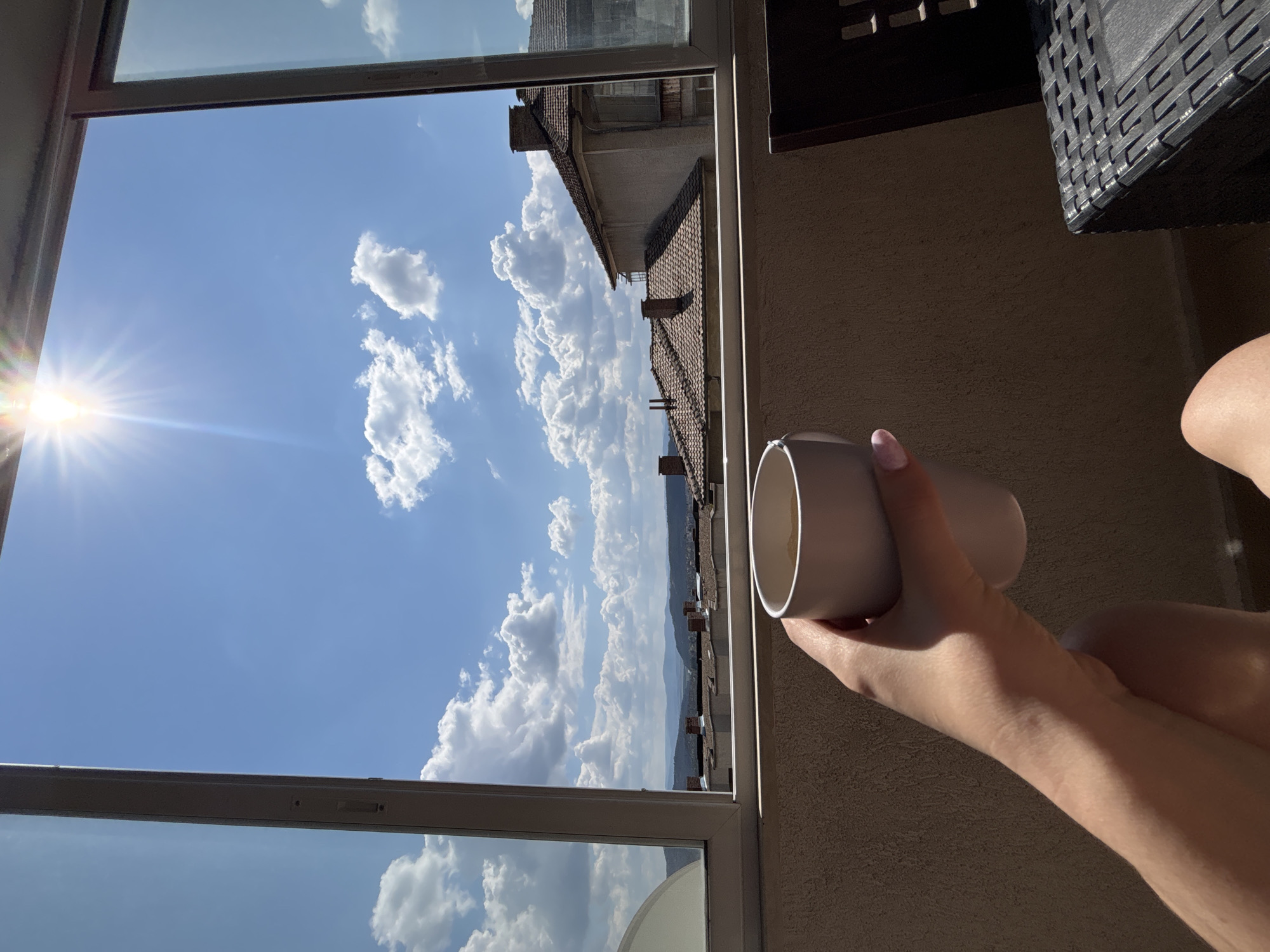 A person is holding a light gray mug with a beige interior, near a window. The window shows a view of Veliko Tarnovo, Bulgaria. The bright sunlight illuminates the left portion of the sky, highlighting the fluffy white clouds. The view beyond the city reveals distant mountains under a clear sky. The window frame is white. Part of a dark, woven table and the person's arm are visible to the right of the window.