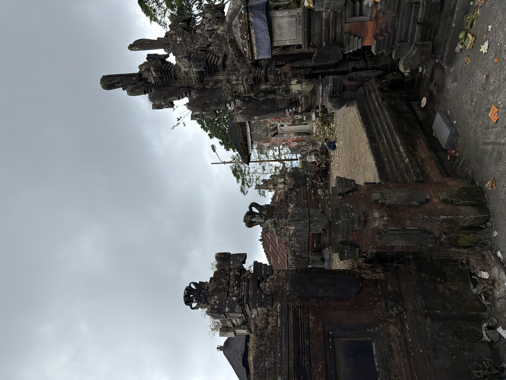 The image depicts the entrance to a traditional Balinese temple in Kintamani, Indonesia, under a cloudy sky. The foreground features weathered, intricately carved stone gateways, adorned with moss and dried vegetation. To the left, a dark, square opening within the gateway suggests an internal structure. Steps lead up to a courtyard area in the middle ground, where several buildings with dark, pitched roofs are visible. One building in the background features a tall, narrow doorway. A person is crouched down in the courtyard, facing away from the camera. The ground is uneven and littered with debris, including plastic bottles and fallen leaves. Green trees and foliage are present in the background, contrasting with the grey sky.