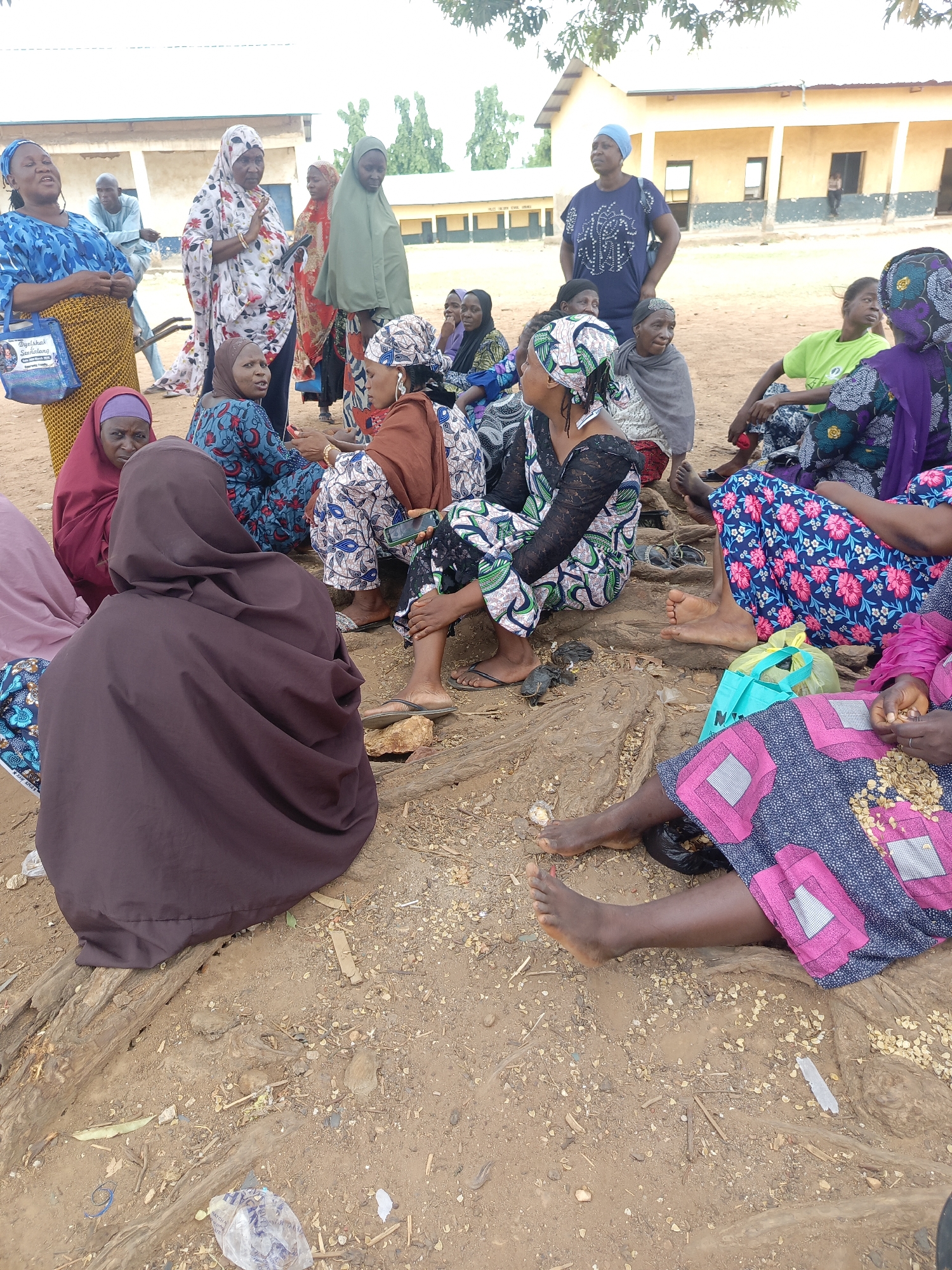 A group of women wearing colorful clothing and head coverings are sitting on the ground in an outdoor area. The ground is dirt. Some women are barefoot. Behind them is a building. Some of the women are sitting on pieces of wood. The location is Akwanga, Nigeria.