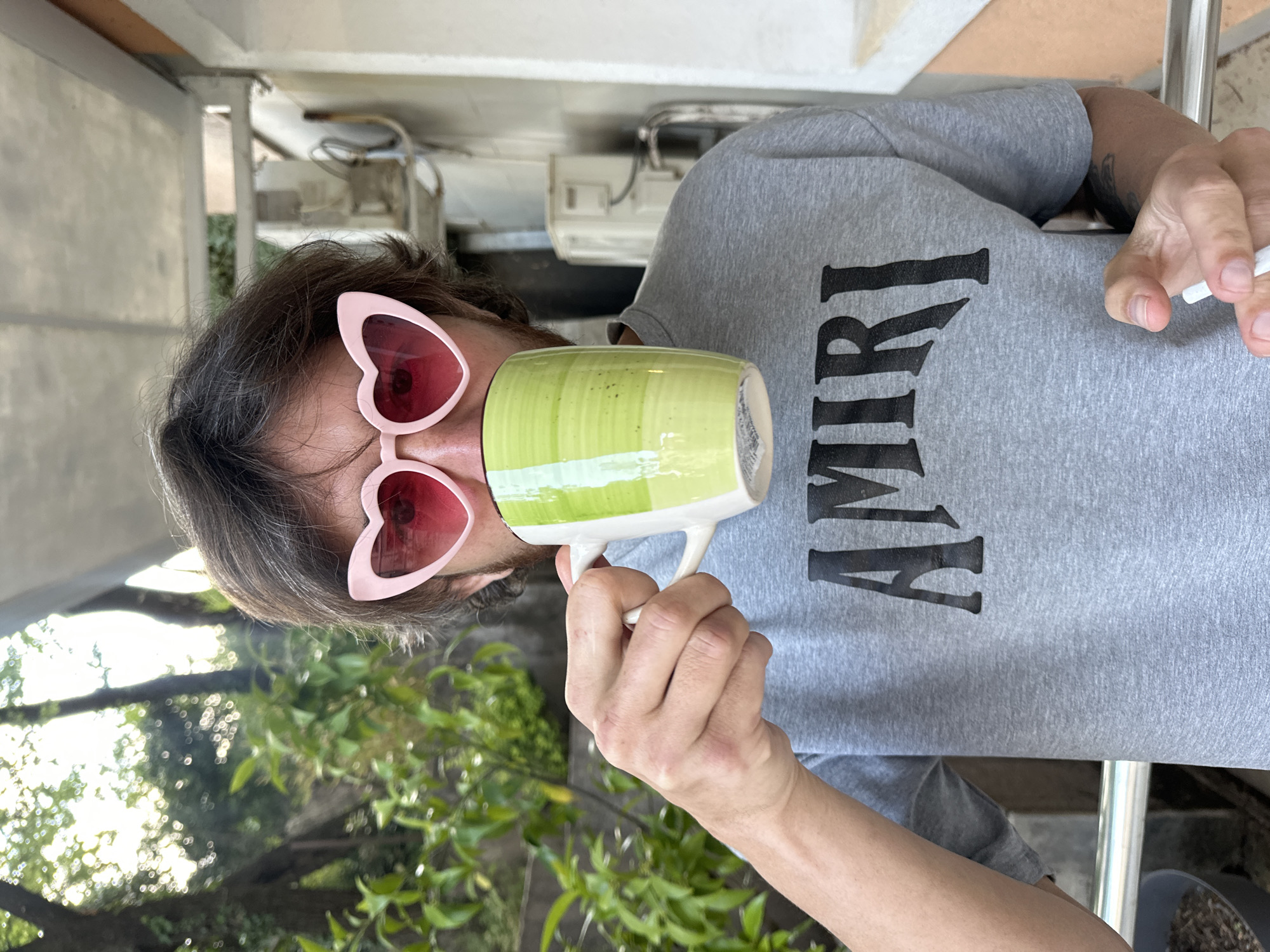 A person wearing pink heart-shaped sunglasses and a gray Amiri t-shirt is holding a green and white mug to their face while smoking a cigarette. The setting appears to be outdoors on a balcony or patio with green foliage visible in the background. The location is Budva, Montenegro.