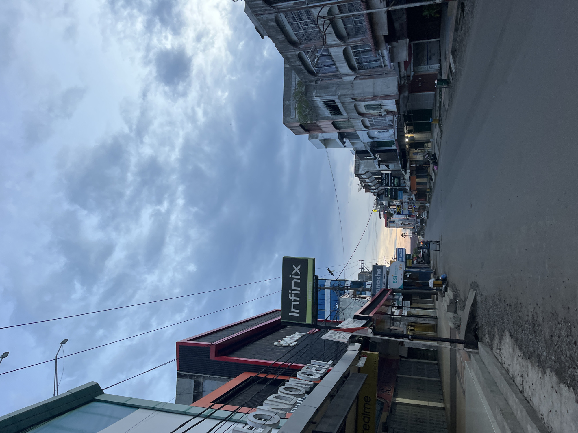 The image shows a street view in Lhokseumawe, Indonesia. The shot is taken at street level, looking down the road. On the left side of the street, there is an Infinix mobile store with prominent branding. Various other shop signs are visible. On the right side of the street, there are older buildings and a sidewalk. The street is paved. The sky is overcast with gray and white clouds. There is no apparent activity visible on the street.