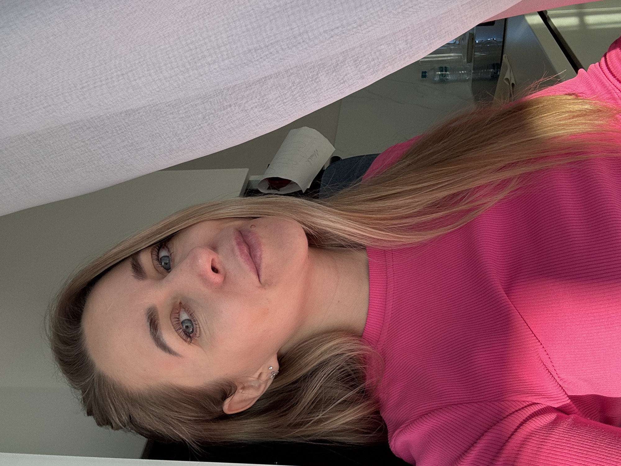 A headshot features a young woman, shown from the chest up, with the image rotated 90 degrees clockwise. She has long, straight blonde hair with lighter highlights, blue eyes, and a fair complexion. She wears a ribbed, long-sleeved top in a vibrant fuchsia or hot pink color. Her gaze is directed towards the camera with a neutral expression, and she has a small, decorative piercing in her left ear.
The setting appears to be indoors, possibly a modern salon or clinic, located in Chaikovsky, Russia. Above her, a large expanse of light-colored, sheer fabric, resembling a curtain or screen, occupies the upper portion of the frame. In the background, behind her head and shoulder, a dark surface resembling a desk or counter is visible. On this surface, a rolled-up piece of white paper can be seen, with faint Russian text "Милый" (Miliy), meaning "Cute" or "Dear," written on it. Further back, out of focus, several transparent bottles, possibly containing water, and reflective surfaces are discernible, suggesting a structured environment. The lighting is bright and even, likely diffused natural light, casting soft shadows and highlighting her hair and the texture of her clothing.