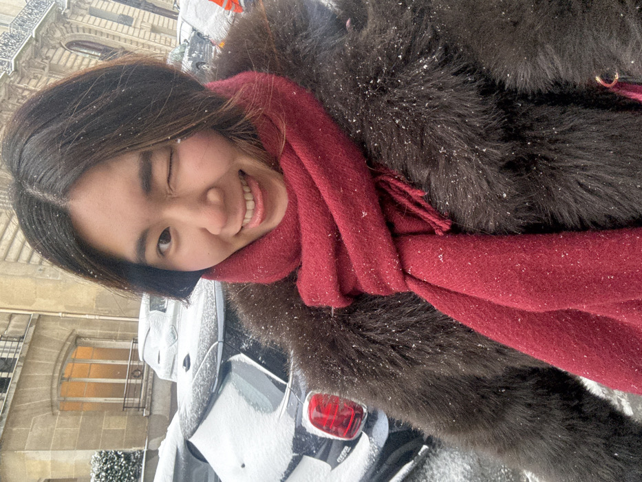 The image depicts a female subject in a snowy urban environment, identified as the city of Paris, France, due to the distinctive Haussmannian architecture visible in the background. The subject, an East Asian woman with dark, chin-length hair, occupies the foreground, looking directly at the camera with her right eye winking and her tongue extended slightly. She is wearing a dark brown, shaggy faux fur coat and a thick, deep red knit scarf wrapped around her neck. Small white flakes, consistent with falling snow, are visible on her hair and clothing. In the background, several parked vehicles, including a dark-colored Mini Cooper partially visible on the left with its red taillight, are covered in a layer of snow. Behind the vehicles, a multi-story, light-colored stone building with ornate architectural details features an arched window emitting warm light. The scene indicates a cold, snowy day.
