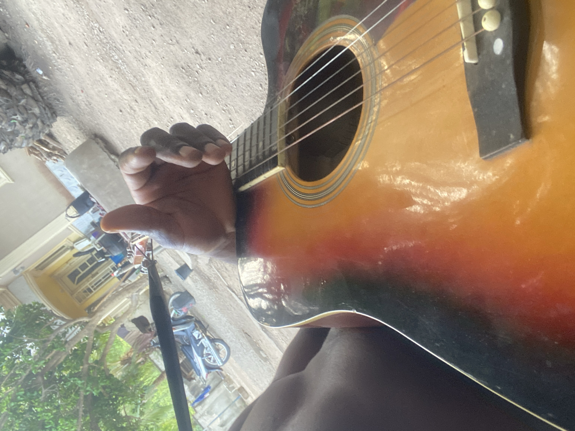 A person with dark skin is holding a guitar in Mararaba, Nigeria. The guitar is an acoustic guitar with a reddish-brown sunburst finish. The person's left hand is gripping the neck of the guitar, while their right hand is out of frame. The background shows a street scene with a house, some trees, and a motorcycle. The image is slightly rotated counter-clockwise.