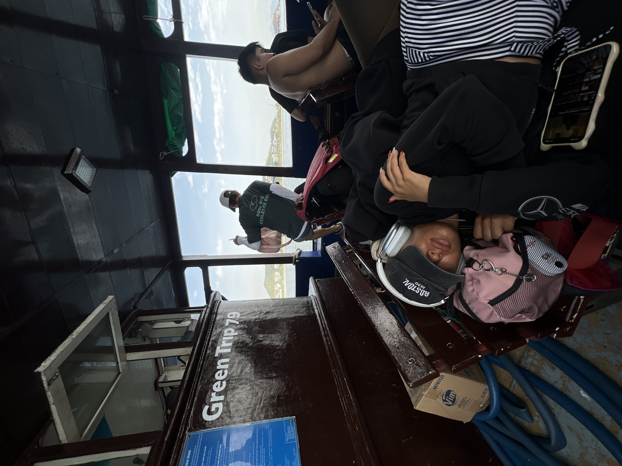 The interior of the "Green Trip 79" boat features several passengers. A young person, wearing headphones and a "Boston 1630" baseball cap, rests their head on a pink backpack. A woman wearing a striped shirt and black pants is beside them, holding a smartphone. A man, shirtless, sits nearby looking at his own phone. A blue hose is coiled on the floor. Through the boat's windows, a glimpse of the shoreline can be seen, placing the scene in Nha Trang, Vietnam.