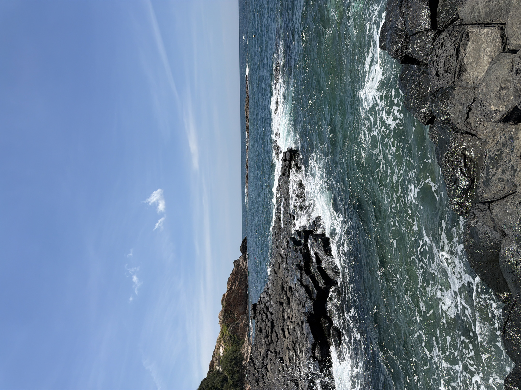 This is a medium shot, eye-level, landscape photograph taken on a sunny day. The image depicts a coastal scene with dark, hexagonal basalt rock formations in the foreground and middle ground, bordering a turbulent, turquoise ocean. Waves crash against the rocks, creating white foam. In the background, a rocky, tree-covered hill slopes down to the water, and a few small boats are visible on the sea. The sky is a clear blue with a few wispy white clouds. The location is identified as Phú Lương, Vietnam, specifically the Gành Đá Đĩa (Stovepipe Rocks) geological formation.