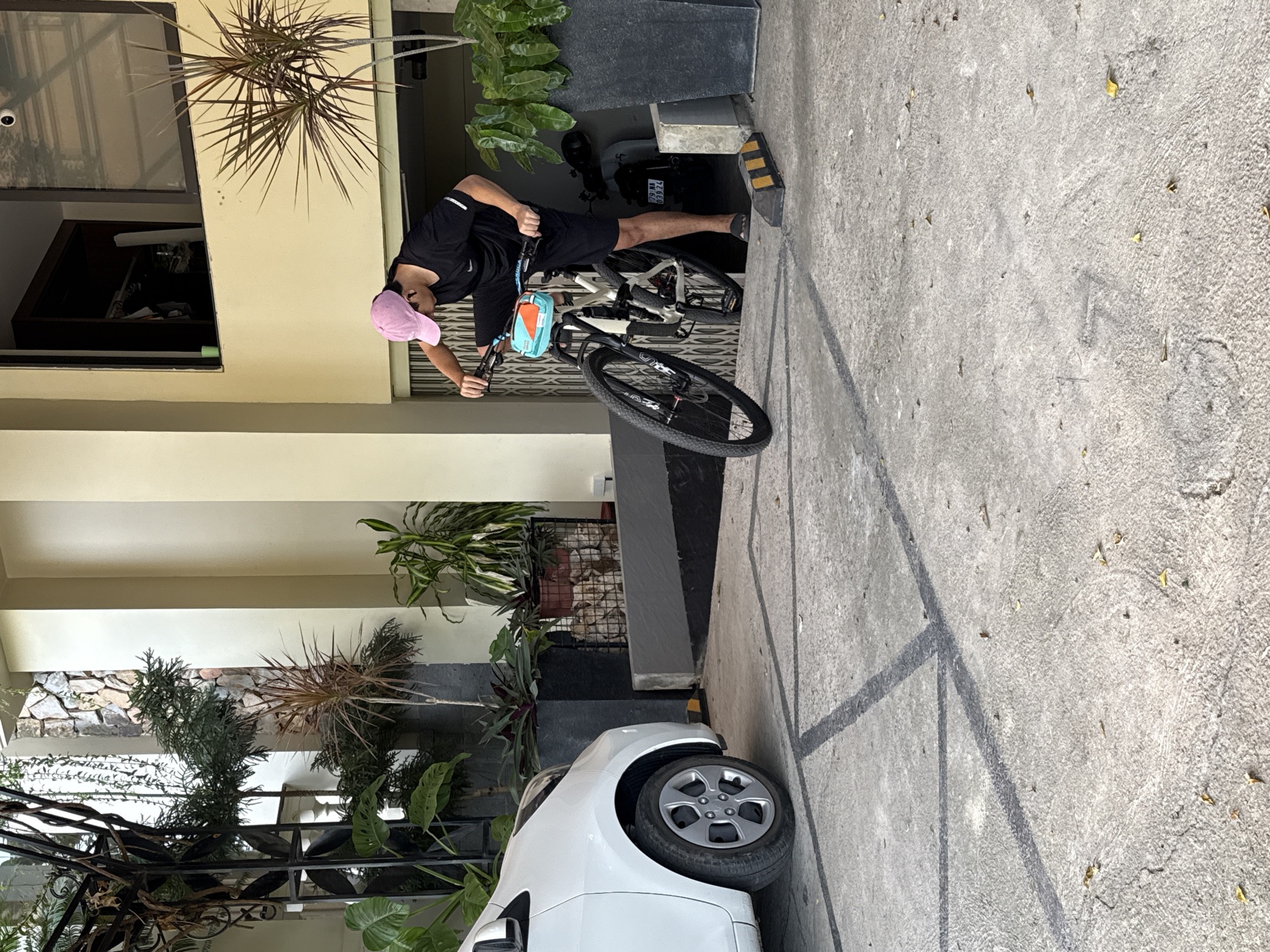 A person wearing a black t-shirt, black shorts, and a pink baseball cap is seen in the image. They are leaning over a white bicycle with a blue and orange bag attached to the handlebars. The person is holding onto the handlebars, suggesting they are either preparing to ride or have just stopped.
The scene appears to be an outdoor setting, possibly a driveway or sidewalk, with a concrete ground marked by dark lines. To the left, there is a building with a pale yellow facade and a window. Plants, including a spiky, dried-looking plant and a leafy green plant, are visible in pots and growing near the building. In the background, a dark structure and some additional greenery are present. A white car is parked in the foreground on the right side of the image.
The lighting suggests it is daytime, with natural light illuminating the scene. The overall impression is one of a casual moment, possibly in a residential or commercial area in Bac Nha Trang Ward, Vietnam, given the location context. There is no discernible text in the image.