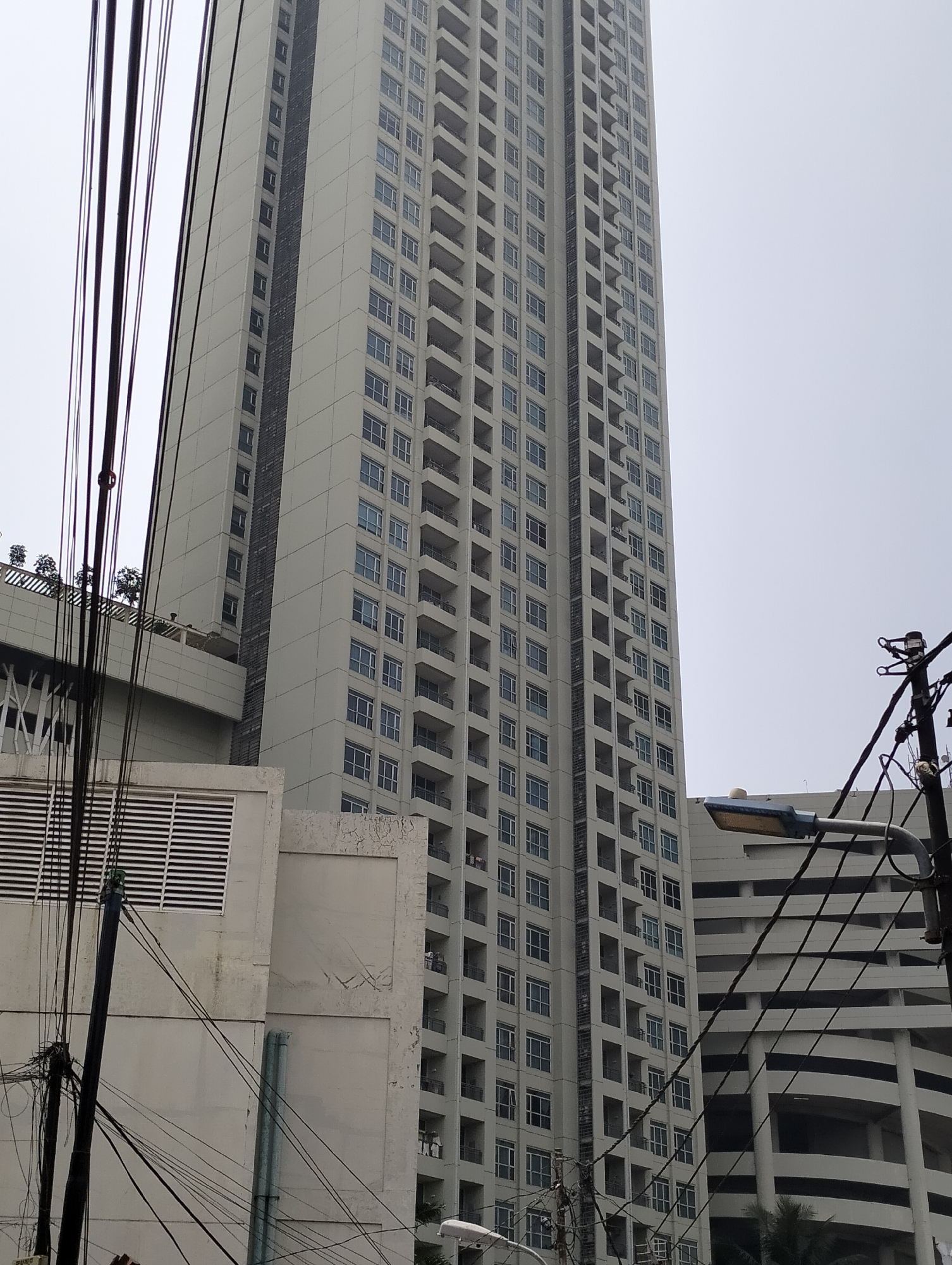 The image depicts a towering, modern residential building in West Jakarta, Indonesia. The building is light-colored with numerous balconies and windows. In the foreground, a dense network of black electrical wires crisscrosses the frame, obscuring parts of the building and a street lamp. To the left, a smaller, white structure with horizontal vents is visible. The sky is overcast and a pale gray. The scene appears to be an urban environment, characterized by dense construction and infrastructure.