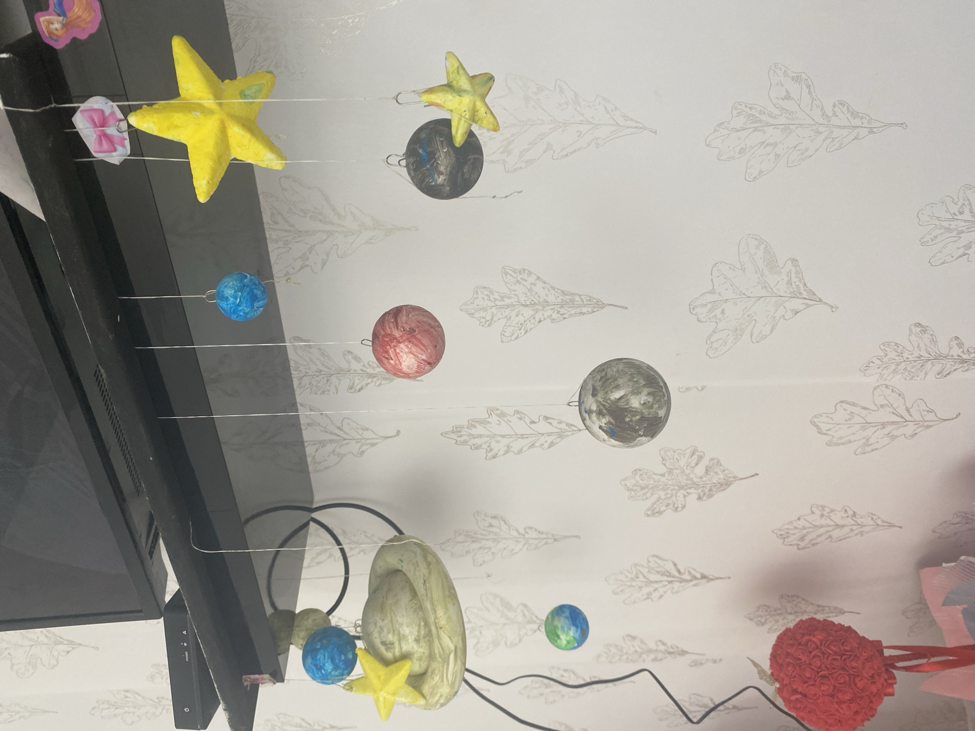 An indoor scene depicts a black shelf mounted on a wall with a repeating oak leaf pattern. A television is partially visible at the top. Hanging from the shelf are several handmade ornaments, including planets and stars, likely part of a mobile. One ornament is a large yellow star, another a pale green ringed planet, and there are smaller spheres in blue, green, and red, along with a gray sphere. A red rose bouquet on a stem is visible in the lower left corner. The setting is not specifically identifiable as being in Blagoevgrad, Bulgaria, based on the visual information provided.