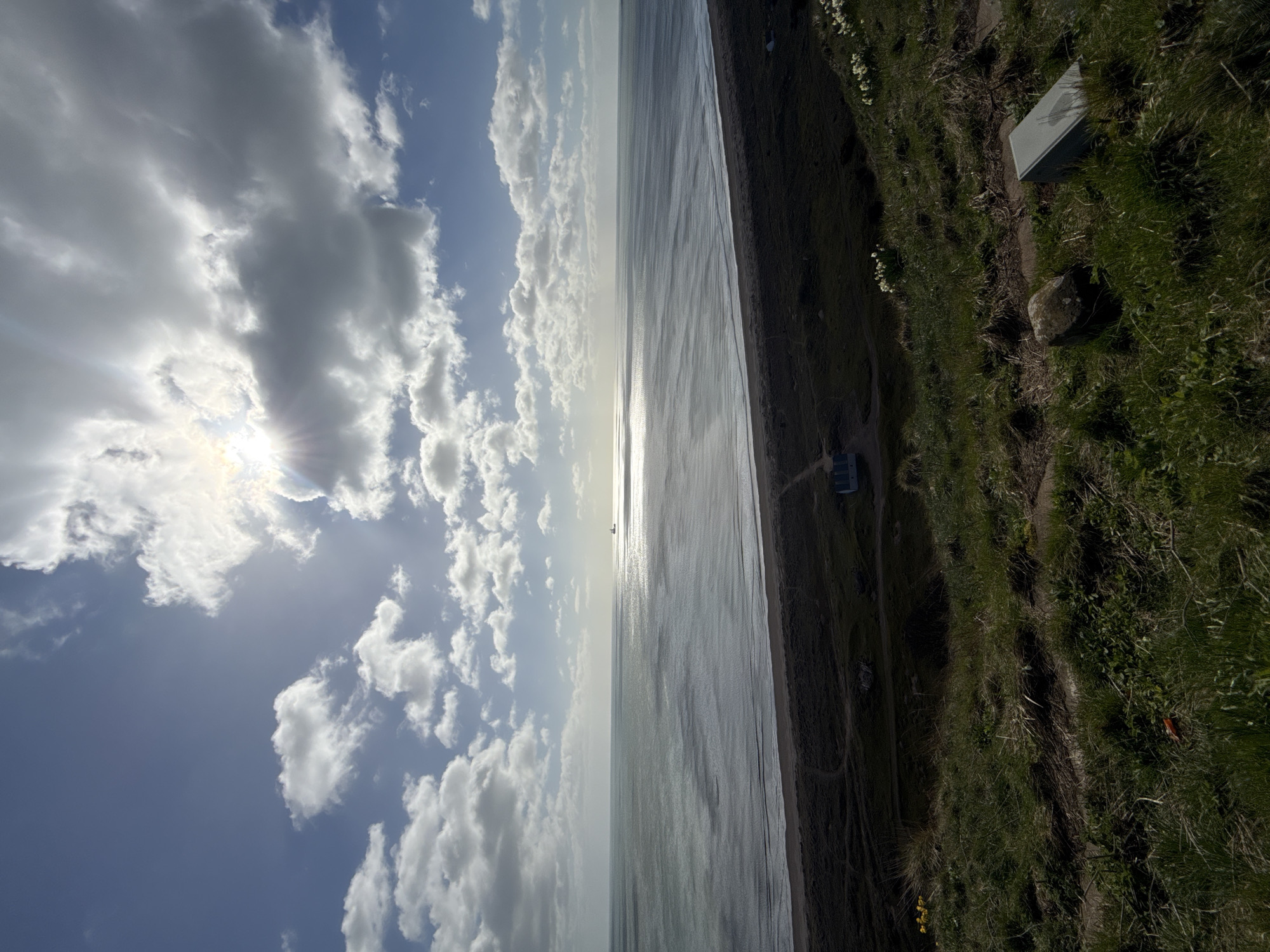 The image shows a view from Montrose, United Kingdom, depicting a shoreline scene. The horizon line divides the image in half, with a cloudy sky above and the North Sea below. The sun is visible through the clouds, creating a bright glare. The sea is calm, with a strip of beach separating it from a grassy coastal area. The grassy area rises slightly, leading to a flat area where a small building and stone structure stand. The image is taken from a high vantage point, and the angle is rotated 90 degrees.