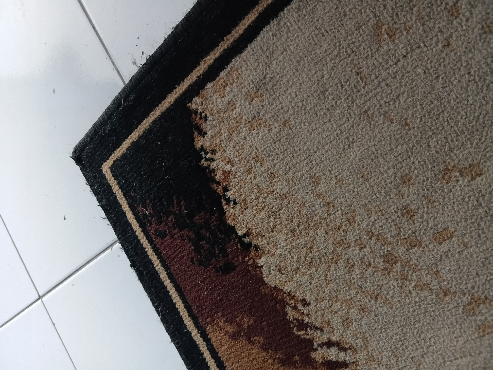 A close-up, high-angle shot shows a carpet with a dark brown, black, and tan abstract pattern lying on a white tiled floor. The carpet's edge is visible in the upper right corner, with a frayed black and tan border. The white tiles are laid in a grid pattern with visible grout lines. The scene is indoors, likely in a residential or commercial space in Tanggul Wetan, Indonesia. No people are present.