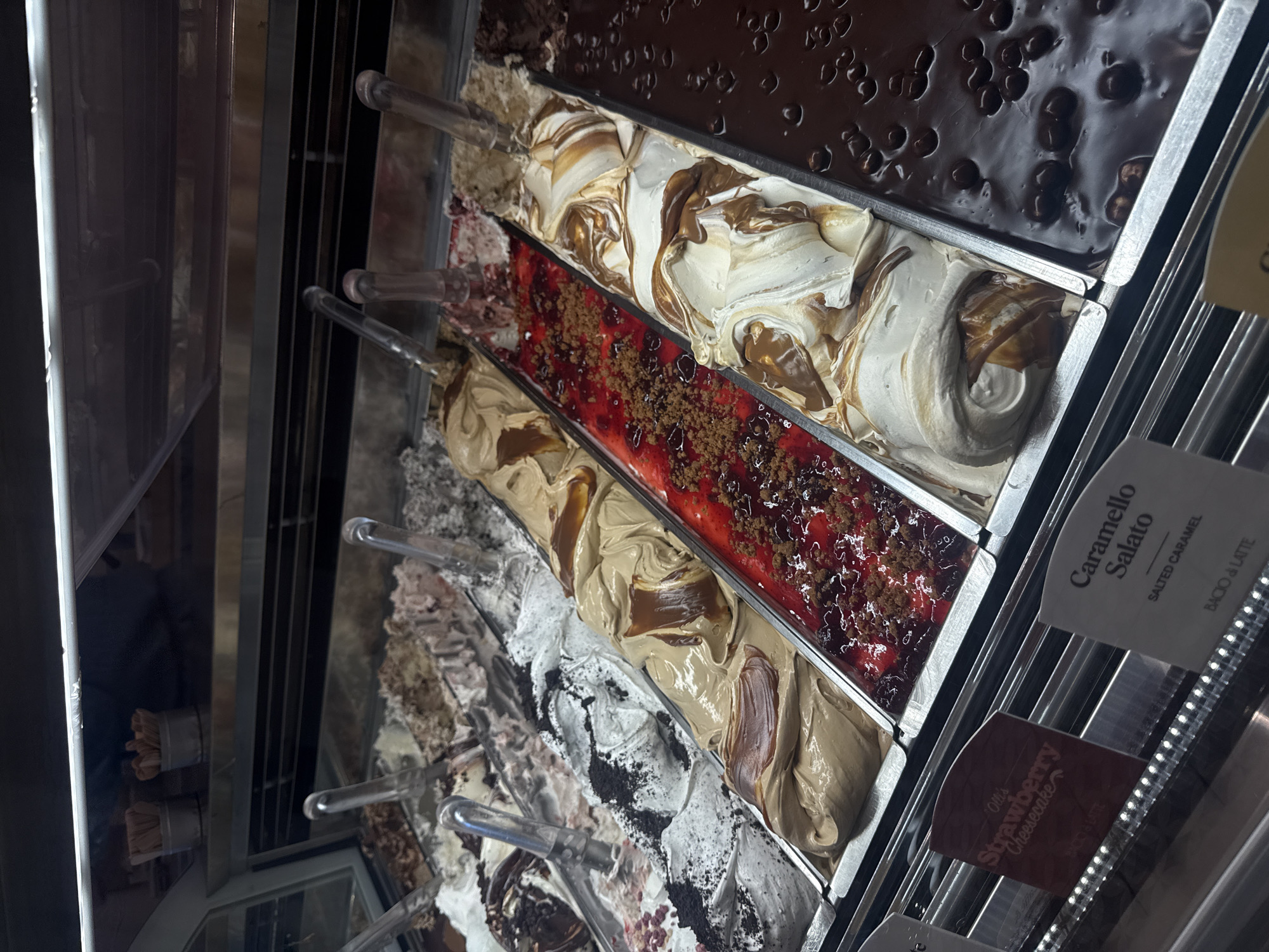 The image shows a display case filled with gelato. The flavors are, from top to bottom: a dark chocolate gelato with dark chocolate pieces, a white gelato with caramel swirl, a strawberry gelato with a streusel topping, a caramel gelato, and cookies and cream. There are labels on the side that say "Caramello Salato, Salted Caramel" and "Strawberry Cheesecake" on the front. It is unknown if the location is in the city of Los Angeles.