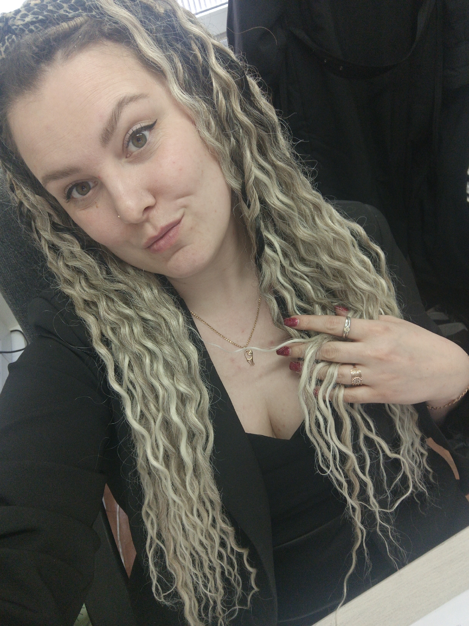 A single female subject is depicted in a close-up, chest-up view, appearing to be an adult. She has long, wavy, blonde hair, with some darker roots visible, styled partly in braids near her forehead, one of which incorporates a leopard-print fabric. Her eyes are light in color, and she wears dark eyeliner and has a small stud piercing on her right nostril. Her lips are lightly pursed. She is wearing a dark-colored blazer or jacket over a dark top. A thin gold-colored necklace with a small pendant is visible around her neck. Her left hand, with red-painted fingernails, is positioned in her hair. Two rings are on her left ring finger: one silver-colored with a yellow stone, and a second gold-colored band. A gold-colored bracelet is on her left wrist. The background is largely dark and indistinct, suggesting an indoor setting. A light-colored surface is partially visible in the lower right corner, consistent with a desk or table. A dark, possibly upholstered object is visible behind her left shoulder. The specific location within Noviy, Russia, or any city, is not discernible from the visual information provided in the image.