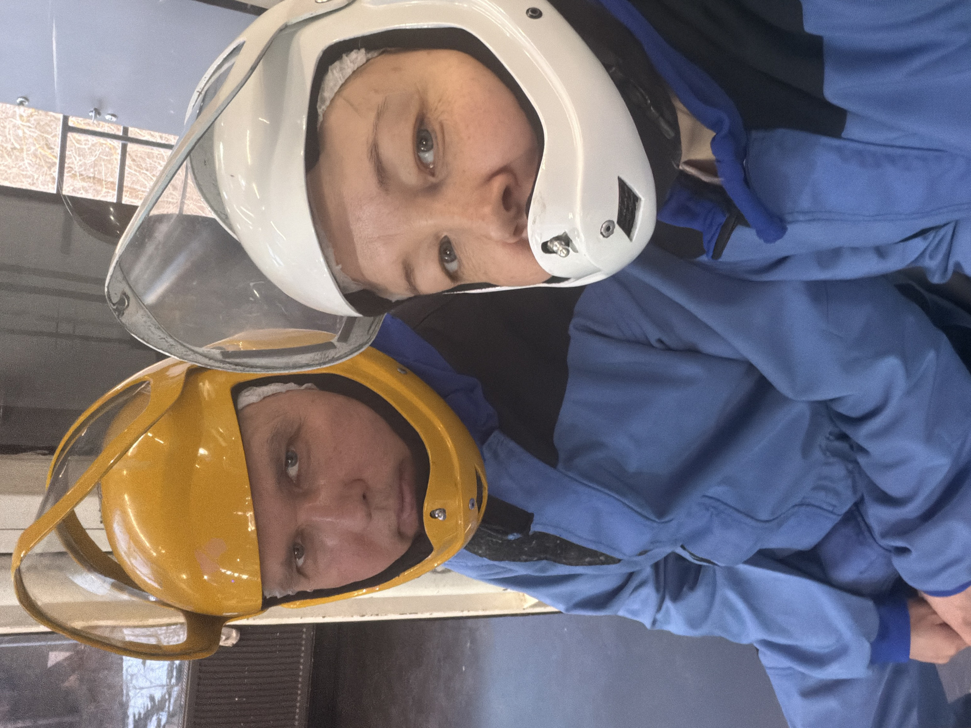 Two people wearing helmets and jumpsuits are posing for a photograph. One helmet is white and the other is yellow, and both have clear visors. The individuals are looking at the camera. The background is blurry and indistinct, but suggests an indoor setting. There is no specific information in the image to confirm the location as Izhevsk, Russia.