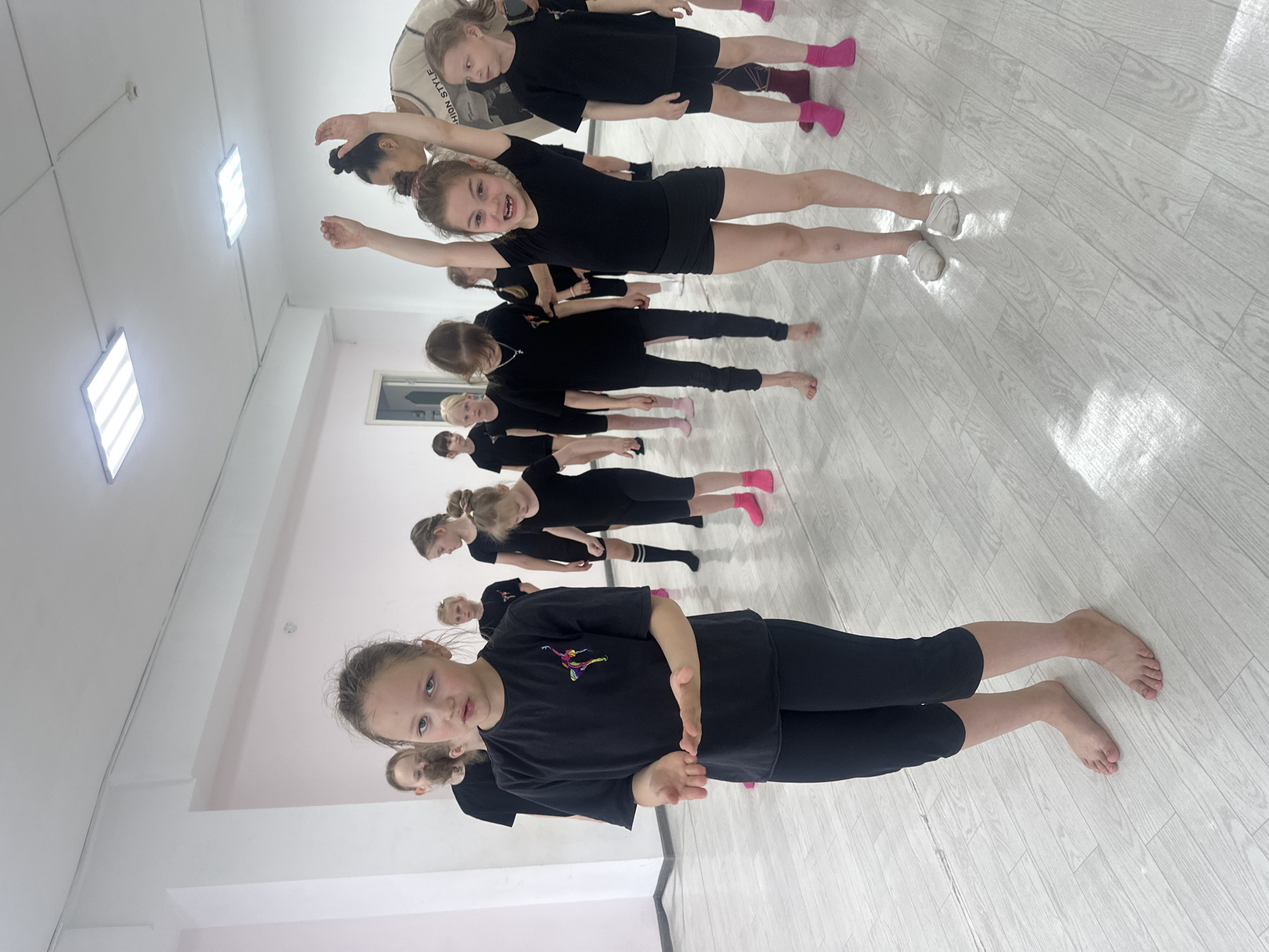 A group of girls in black dance attire is practicing in a studio with a light wood floor. The studio has white walls and a ceiling with recessed lighting fixtures. There is a single window on the wall. One girl is in the foreground, kneeling, while others are lined up behind her, some standing with arms raised. Most of the girls are barefoot, while a few wear pink socks or white dance shoes. The picture gives no indication of the location in Votkinsk, Russia.