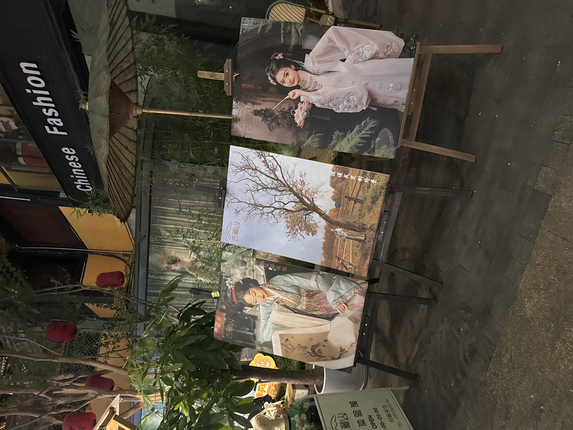 In Yuzhong District, China, three large printed artworks are displayed on easels on a paved walkway. The artworks feature portraits of young women in traditional Chinese attire. The first artwork, on the left, shows a woman in a light blue and pink hanfu, holding a scroll. The middle artwork depicts a landscape scene with a barren tree and a woman in the distance. The rightmost artwork features a woman in a flowing pink hanfu, holding a lotus flower. Behind the artworks, there is a store with a sign that reads "Chinese Fashion". Red lanterns are visible hanging from trees in the background, and a traditional umbrella is also present. The scene appears to be an outdoor display, possibly in a shopping area or cultural district, set up at dusk or night, as indicated by the artificial lighting and the dark sky.