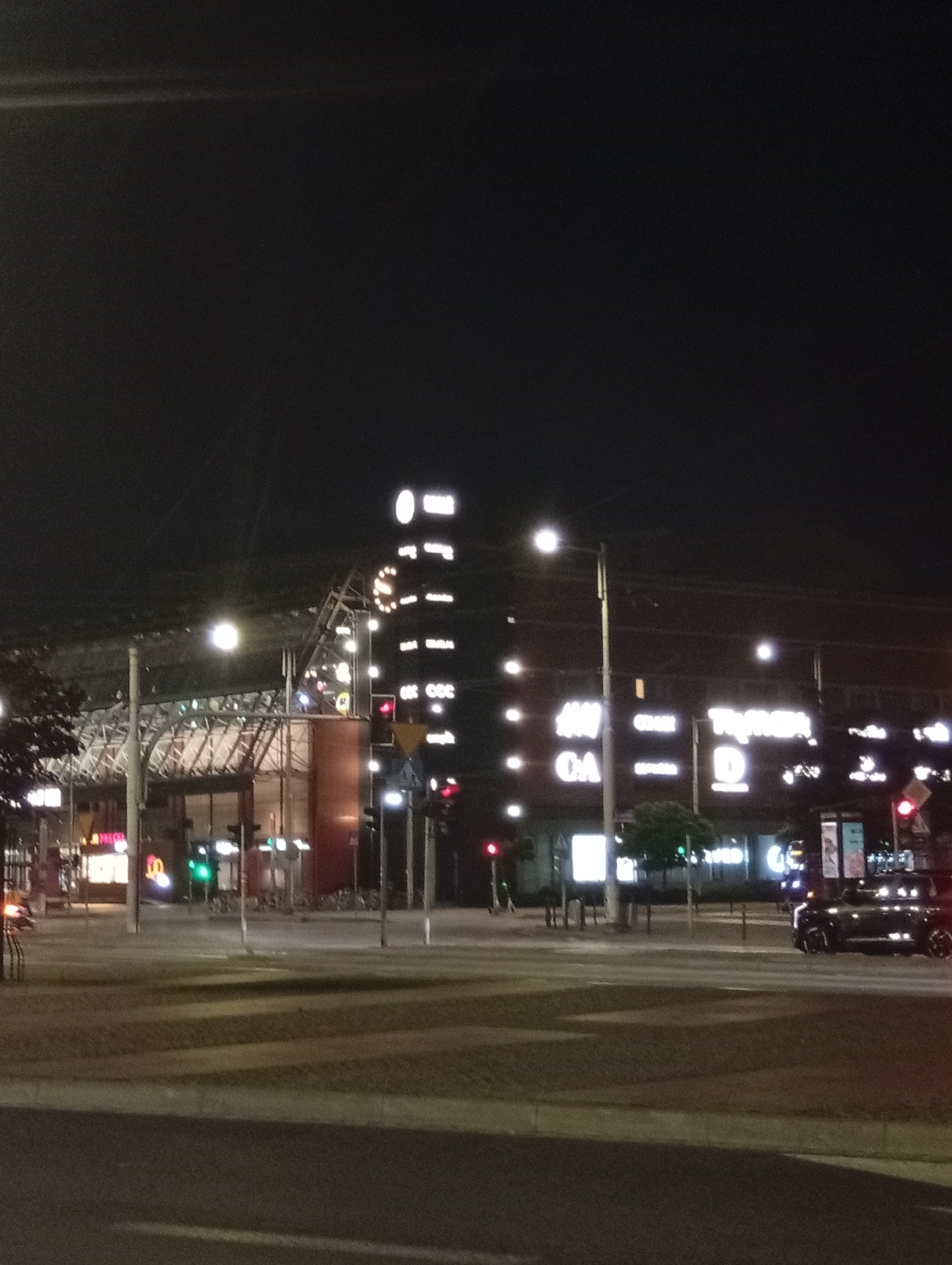 This is a nighttime, eye-level, medium shot of a street in Wrocław, Poland. The scene is dominated by a large building with illuminated signage for various retail stores, including H&M, CA, and T-Mobile. The building appears to be part of a shopping center or a complex with multiple storefronts. A black SUV is visible on the right side of the image, in motion or stopped at an intersection. Traffic lights are present, with some showing red. The street is paved, and a crosswalk is visible. Trees and lampposts are also part of the environment. The overall atmosphere is urban and commercial, with the bright lights of the stores contrasting with the dark night sky.