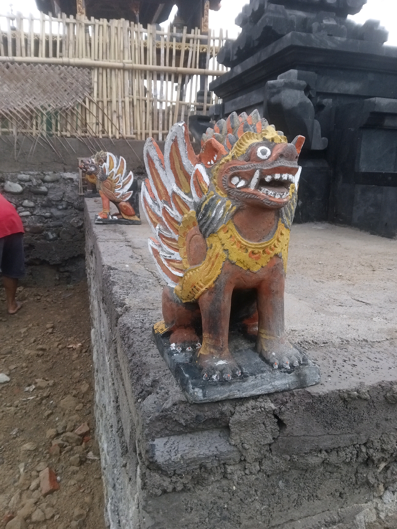 In Mendoyo, Indonesia, a weathered stone wall serves as a platform for two vibrantly painted statues of mythical creatures, likely guardians. The statue in the foreground, a fierce, lion-like being with orange, yellow, and white detailing, is positioned on the right, facing left with its mouth agape, revealing sharp teeth. Its elaborate feathered wings are prominently displayed. To the left and further back, a similar statue, though partially obscured, also faces left. Behind the statues, a dark, stone structure, possibly a temple entrance, rises in the background. A bamboo fence, constructed from vertical poles and woven matting, stretches across the middle ground, suggesting a boundary or enclosure. The ground in front of the wall is uneven, with exposed soil, small stones, and scattered red bricks, indicating ongoing construction or renovation. To the far left, a person wearing a red shirt and dark shorts stands partially in frame, their legs and feet visible, suggesting their presence in the area. The overall scene conveys a sense of cultural heritage and active restoration within a rural Indonesian setting.
