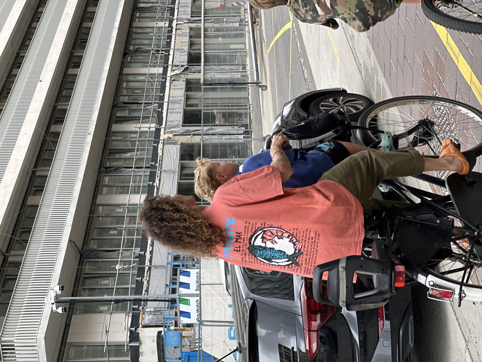 In Amsterdam, Netherlands, a woman with curly brown hair rides a bicycle with a young boy seated behind her. The woman wears an orange t-shirt with a graphic design of a fish and shrimp, and khaki pants. The boy wears a blue shirt and black shorts. They are positioned on a paved street, with a dark-colored car to their left. In the background, a multi-story building with balconies and numerous windows is under construction, with scaffolding visible. The scene appears to be on a cloudy day.