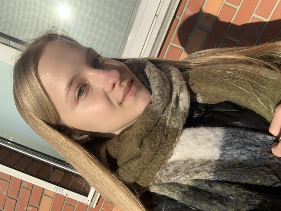 The image depicts a close-up portrait of a young female with long, straight, blonde hair, light-colored eyes, and fair skin, showing a slight smile. She is wearing a thick, multi-tonal knit scarf in shades of olive green, white, and dark blue/grey, wrapped around her neck over a dark-colored jacket. A white wireless earbud is inserted into her right ear, accompanied by a small silver hoop earring. The background consists of a red brick wall on the left and lower right, and a white-framed window above, prominently reflecting direct sunlight. Strong natural light illuminates the subject's face and hair, and her shadow is cast onto the adjacent brick wall. No specific architectural landmarks or signs are visible to identify a precise location within Kyiv, Ukraine.
