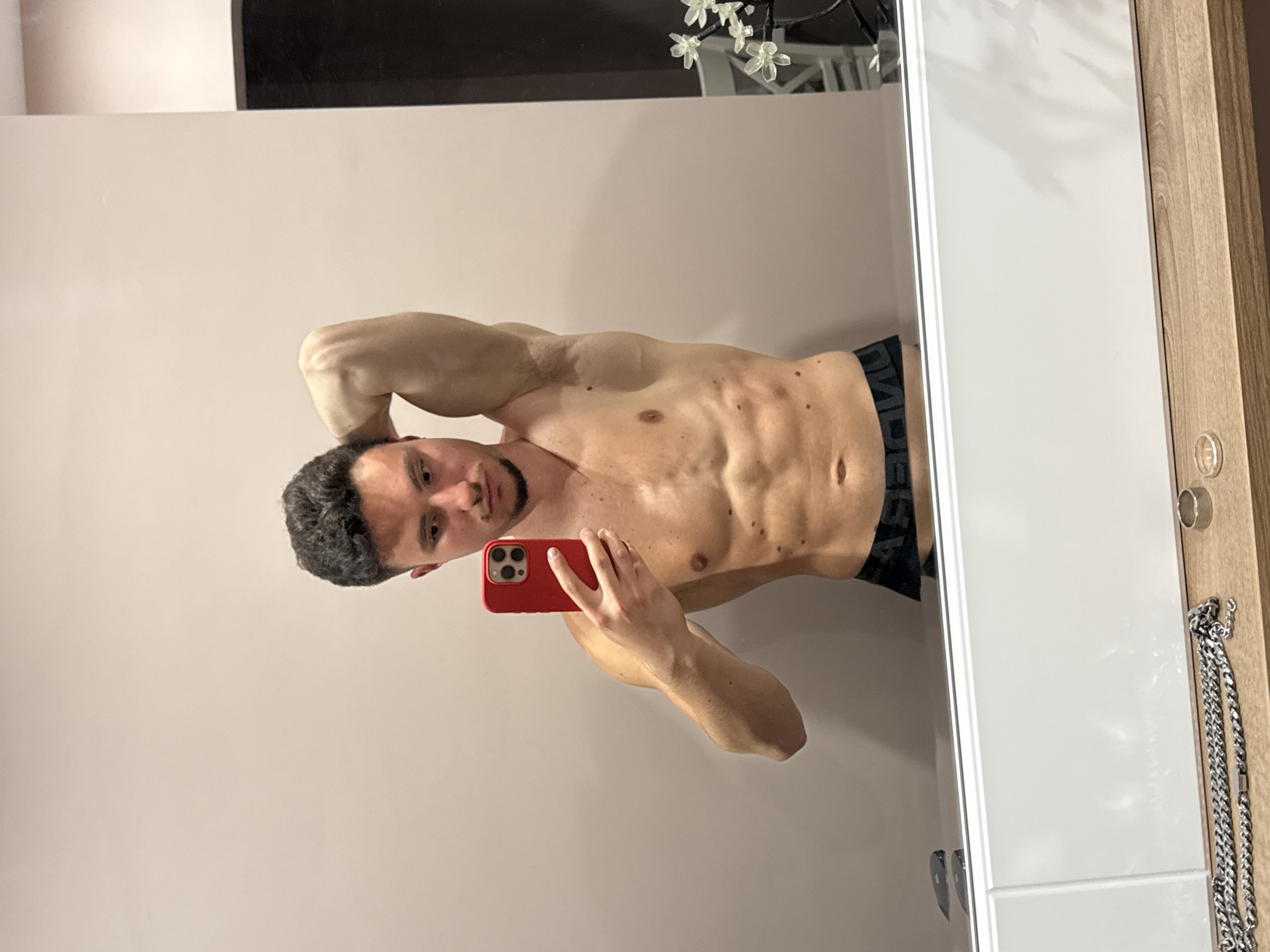 A shirtless man is taking a selfie in a mirror. He holds a red smartphone in his right hand. He is wearing dark underwear with a visible logo. The background consists of a beige wall and a white cabinet door. A silver necklace is hanging on the side of the cabinet. There is no clear evidence to determine the location in the city of Sofia, Bulgaria.