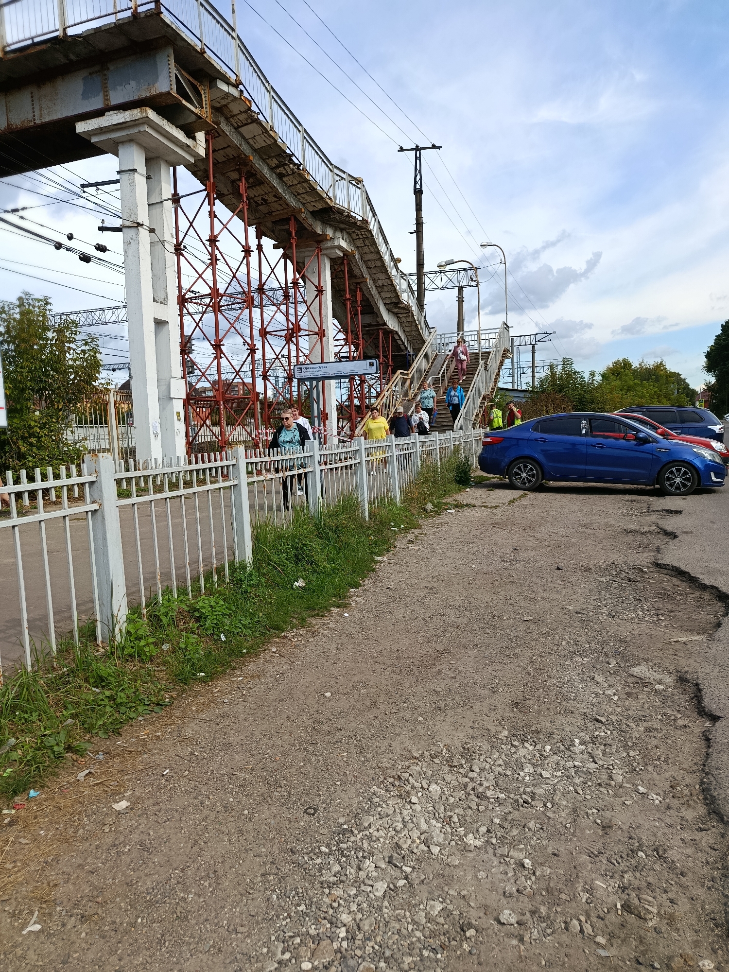 The image depicts a pedestrian overpass at a railway station in Orekhovo-Zuyevo, Russia. A blue sedan is parked to the right of the scene, near a dilapidated asphalt road. Several people are seen on the overpass, ascending and descending the stairs. The overpass itself is a substantial structure, supported by concrete pillars and a lattice of red metal bracing. A white metal fence runs along the side of the walkway leading to the overpass, bordered by overgrown grass. Overhead, power lines and utility poles are visible against a cloudy sky. The overall impression is of a functional but aging public infrastructure in an outdoor urban environment.