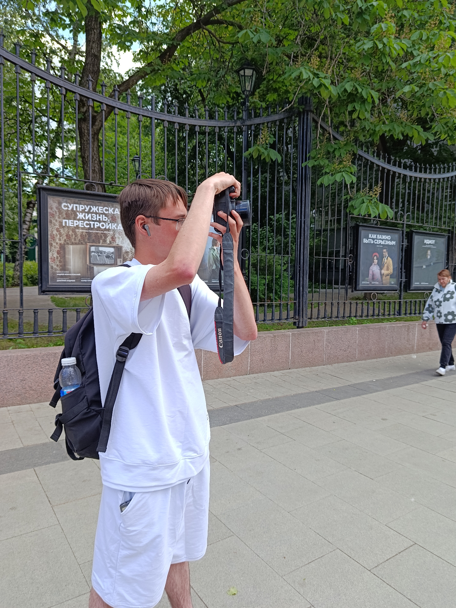 A young man is captured mid-action, taking a photograph outdoors in what appears to be Moscow, Russia. He stands on a light-colored paved sidewalk, holding a black DSLR camera with a "Canon" branded strap up to his eyes, focusing intently. He is dressed in a white short-sleeved t-shirt and white shorts, wearing glasses and earbuds. A black backpack, containing a clear plastic water bottle in a side pocket, is slung over his left shoulder.
Behind the man, a tall, ornate black metal fence separates the public walkway from a verdant area filled with trees and foliage. Several framed informational or exhibition posters are visible through the fence. The most prominent poster to the man's left features Russian text: "СУПРУЖЕСКАЯ ЖИЗНЬ. ПЕРЕСТРОЙКА" (MARRIED LIFE. PERESTROIKA), accompanied by smaller text and an image of an old television. Further to the right along the fence, another poster displays the text "КАК ВАЖНО БЫТЬ СЕРЬЕЗНЫМ" (HOW IMPORTANT IT IS TO BE SERIOUS), and a third reads "ИДИОТ" (IDIOT), both featuring artistic depictions.
On the right side of the image, a woman walks away from the viewer on the same sidewalk. She wears a light-colored, floral-patterned jacket and dark trousers. The scene is illuminated by natural daylight, suggesting a pleasant outdoor setting during the day, possibly in a park or public square. The trees are lush green, indicating a temperate season.